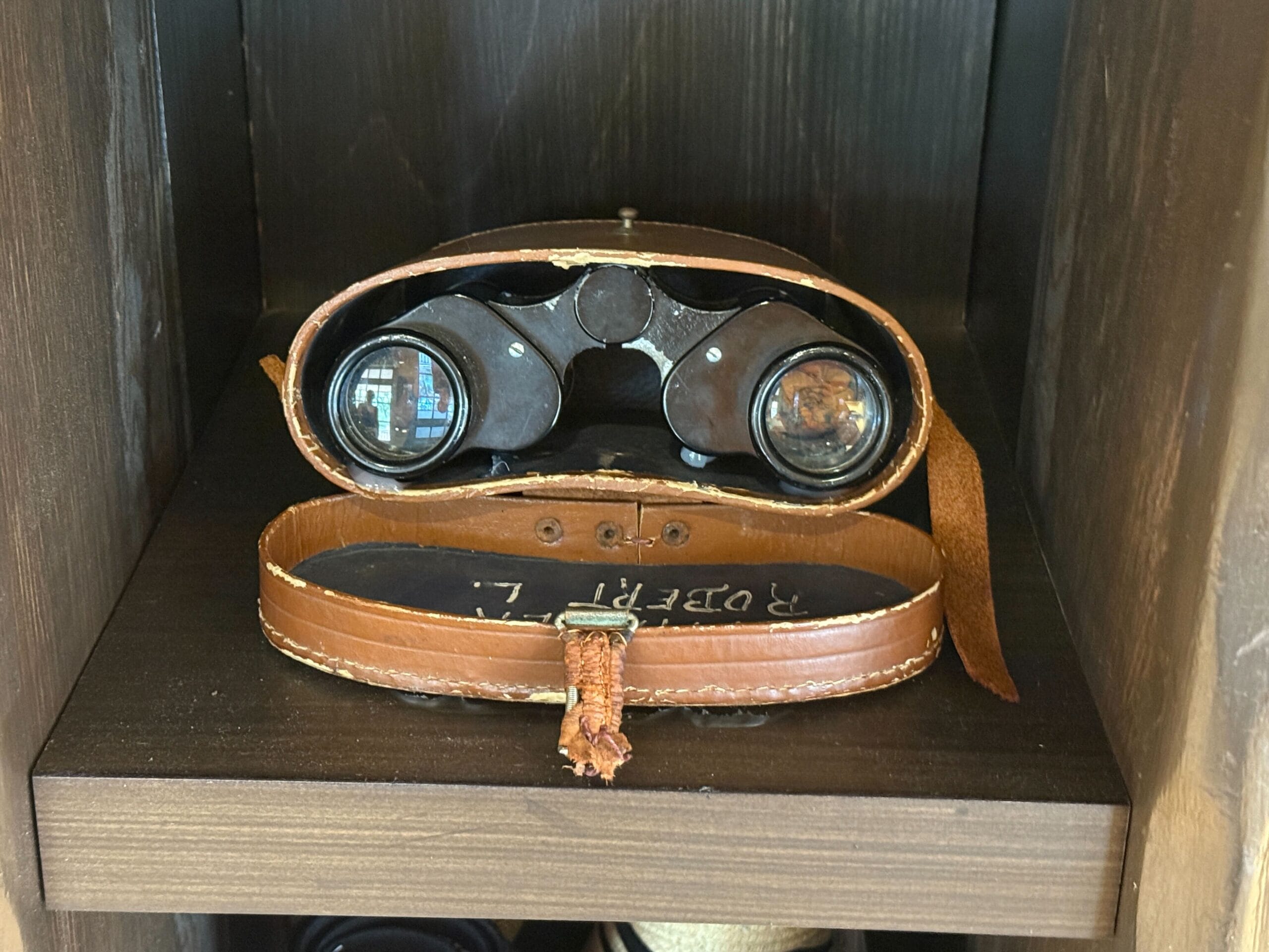 A pair of vintage binoculars in a brown leather case sits on a Prairie Outpost shelf.