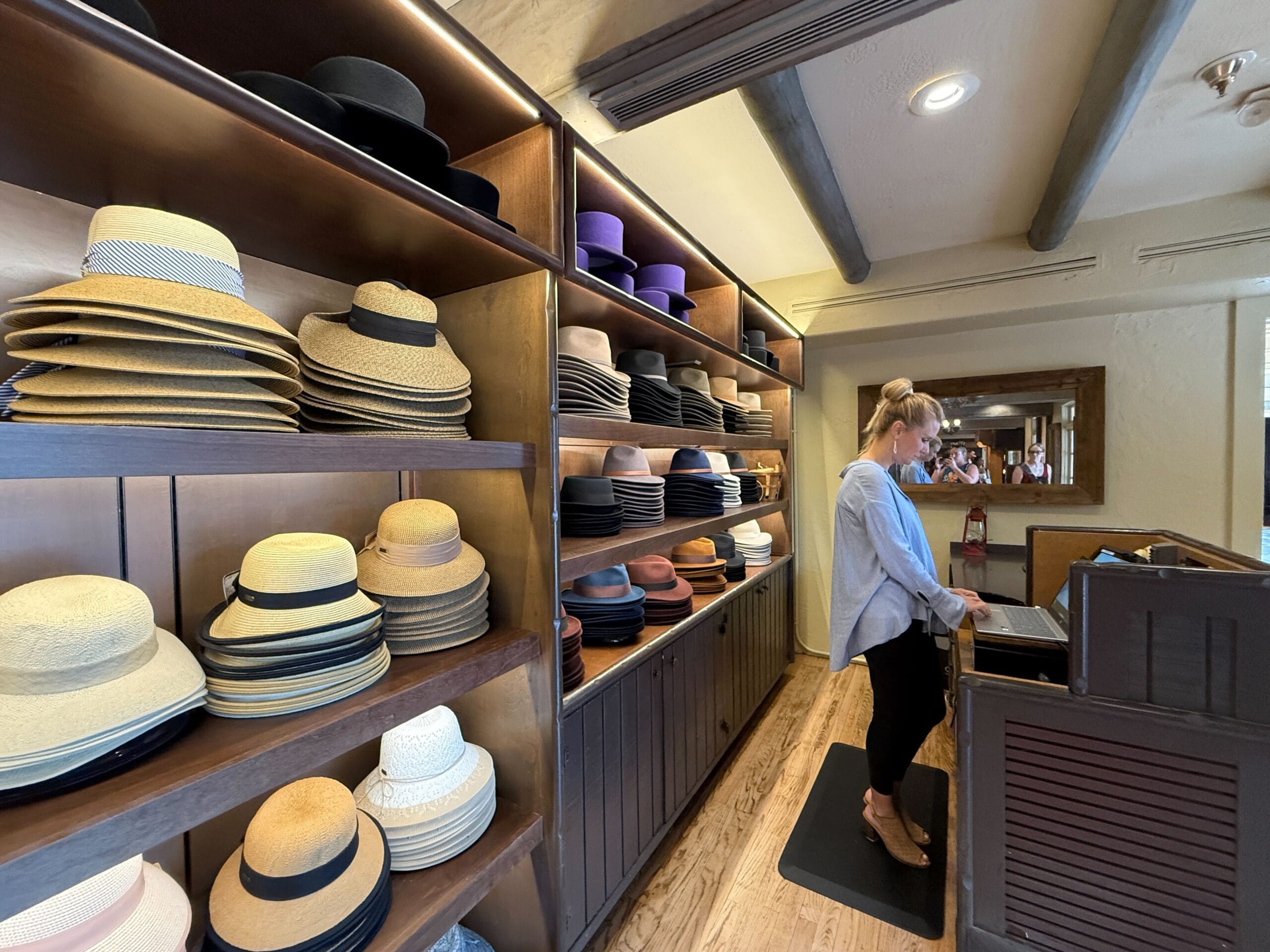 A woman stands at the counter of Prairie Outpost, a charming hat shop with neatly stacked hats on wooden shelves.