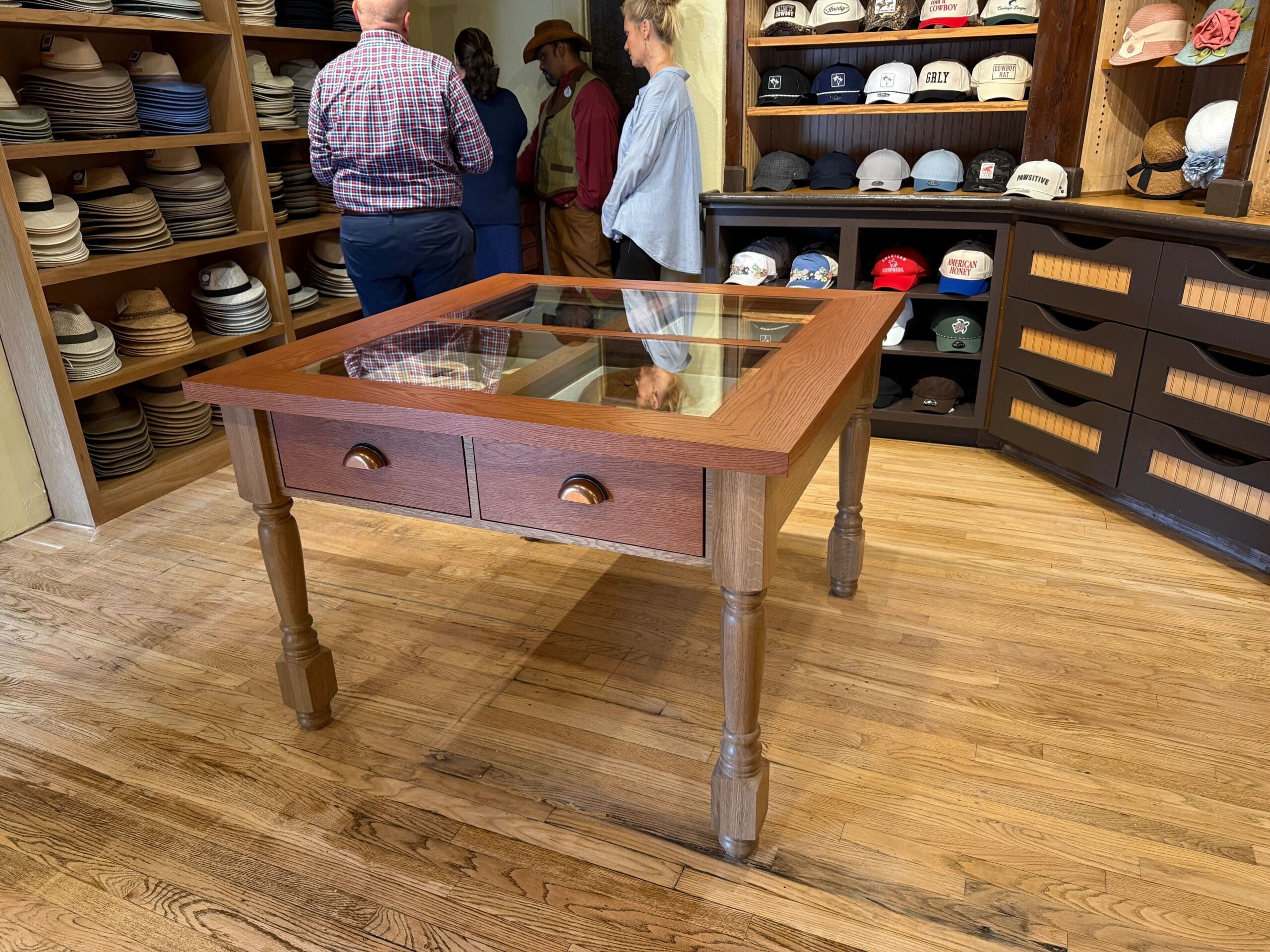 A wooden display table with glass top in a Prairie Outpost Hat Shop, hats on shelves and people talking in the background.