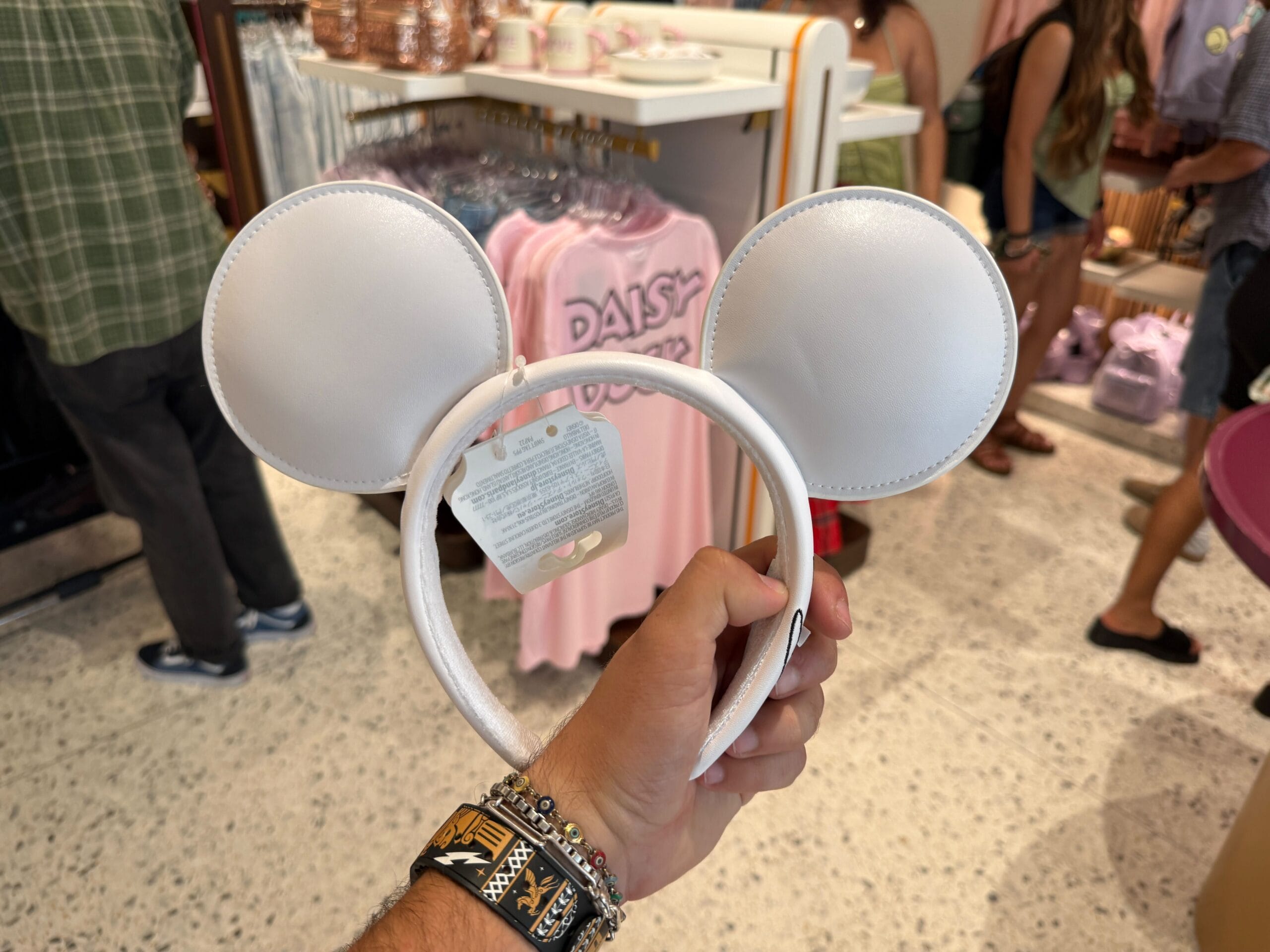 A hand holds white Mickey Mouse ears headband at Disneyland Resort, with pink Daisy Duck shirts in the background.