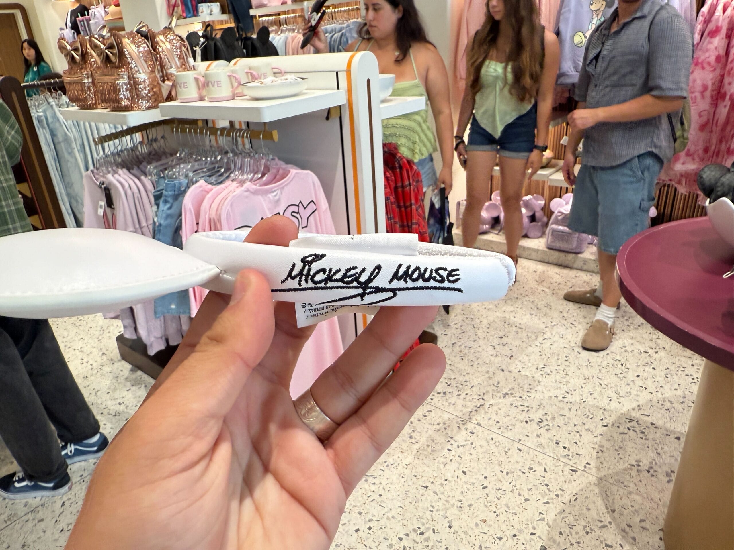 A hand holds a white Disneyland Resort headband labeled "Mickey Mouse" in a busy store with clothing and plush items.