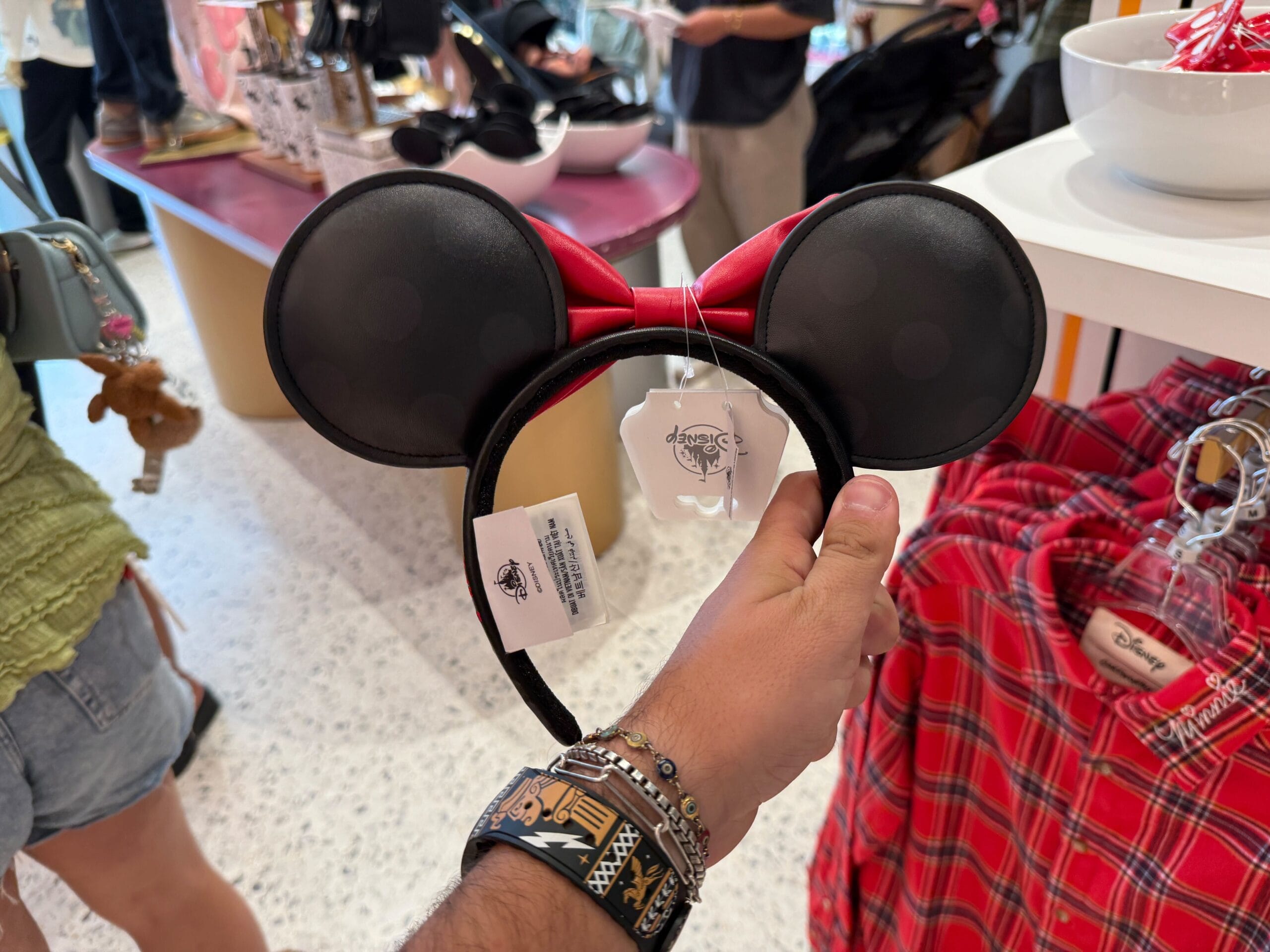 A hand holds a Minnie Mouse ears headband with red bow at Disneyland Resort, beside red plaid shirts.