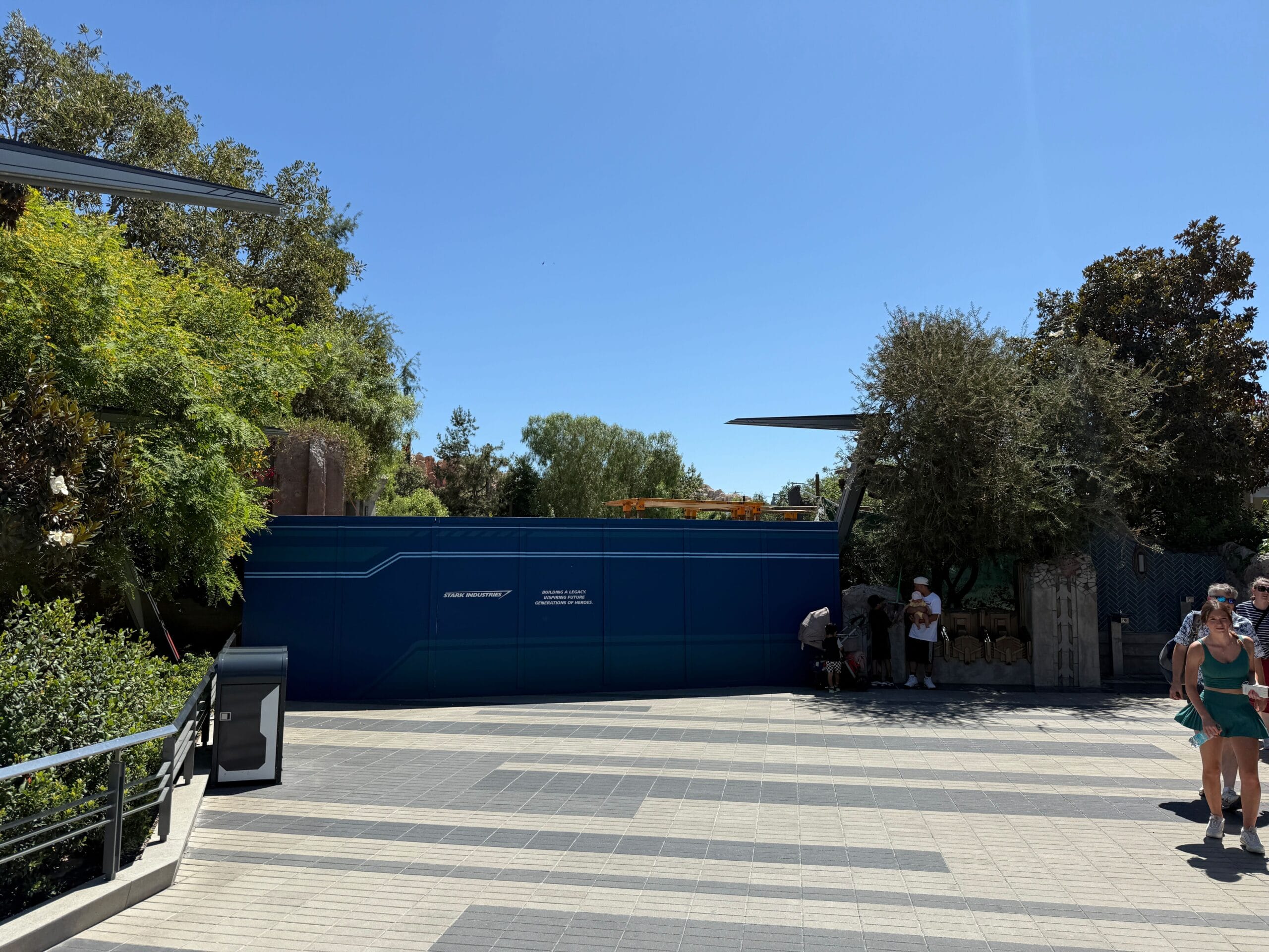 A blue construction wall for Avengers Campus Expansion with people walking nearby, surrounded by trees under a clear blue sky.