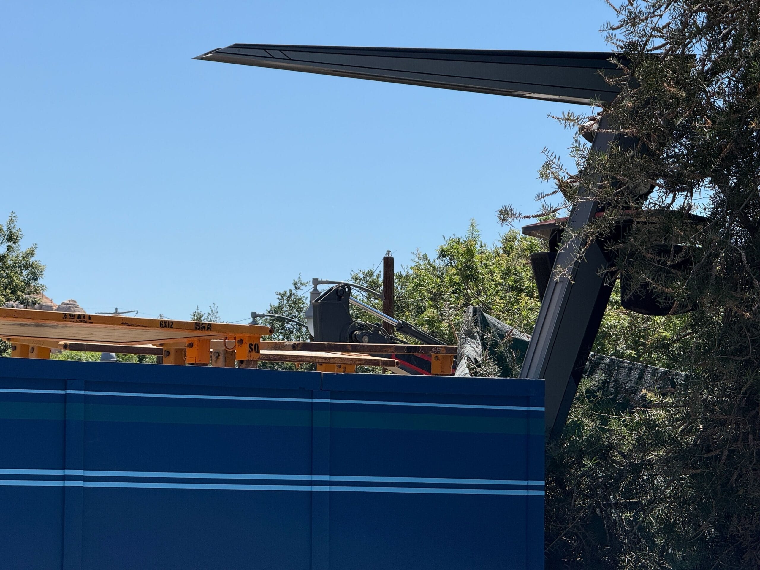 A large, black metal structure rises behind a blue construction barrier and trees under a clear sky.