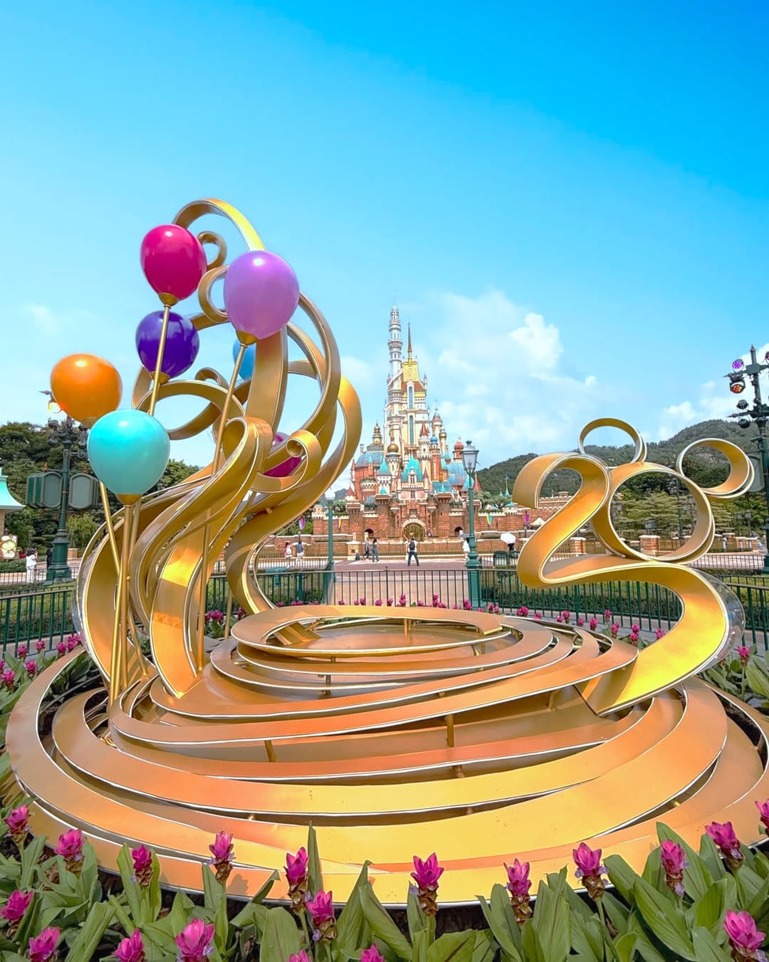 Golden "100" sculpture with colorful balloons in front of a fairytale castle, surrounded by flowers and blue sky.