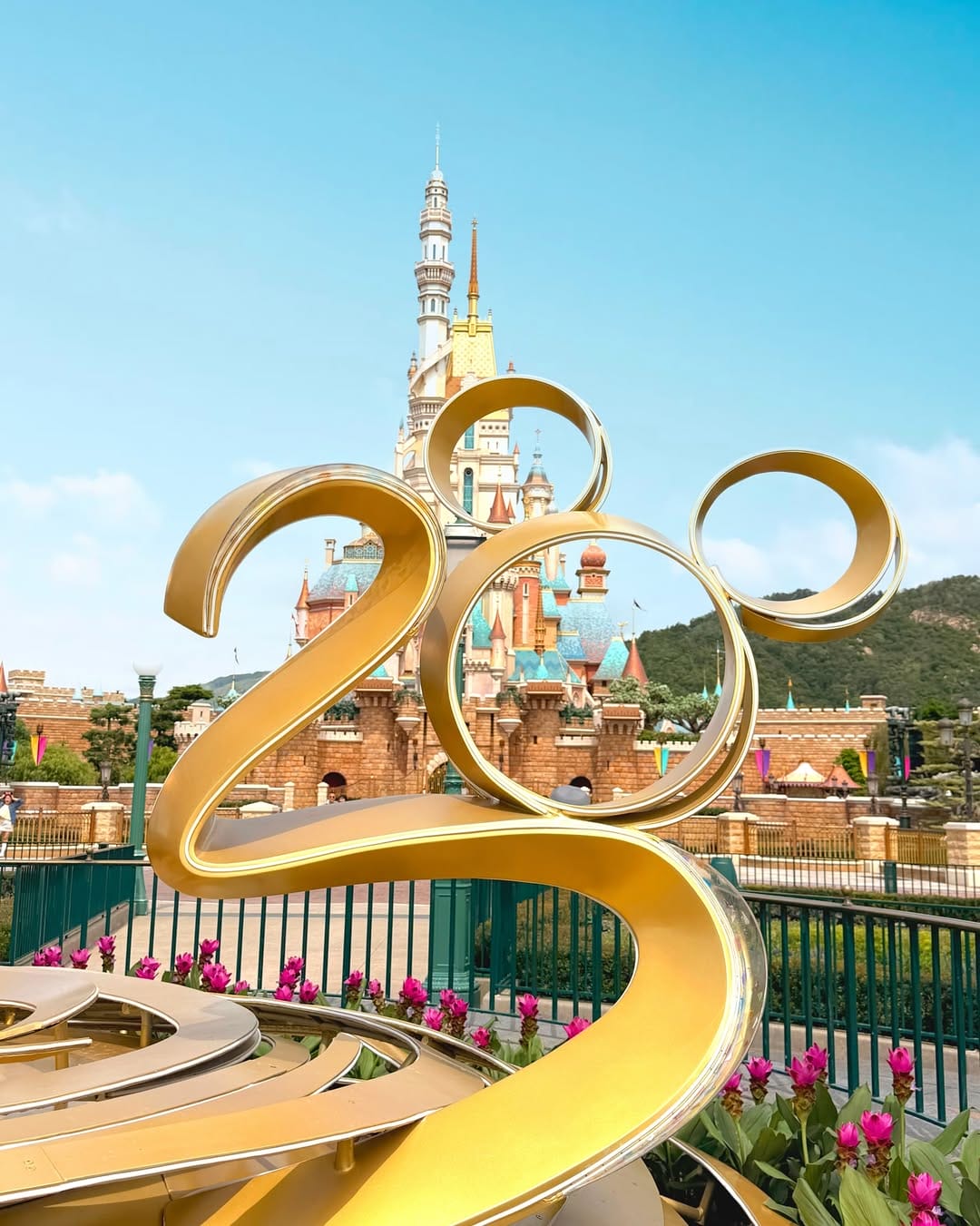 Golden "25" statue with Mickey Mouse ears in front of a fairytale castle at a theme park, under a clear blue sky.