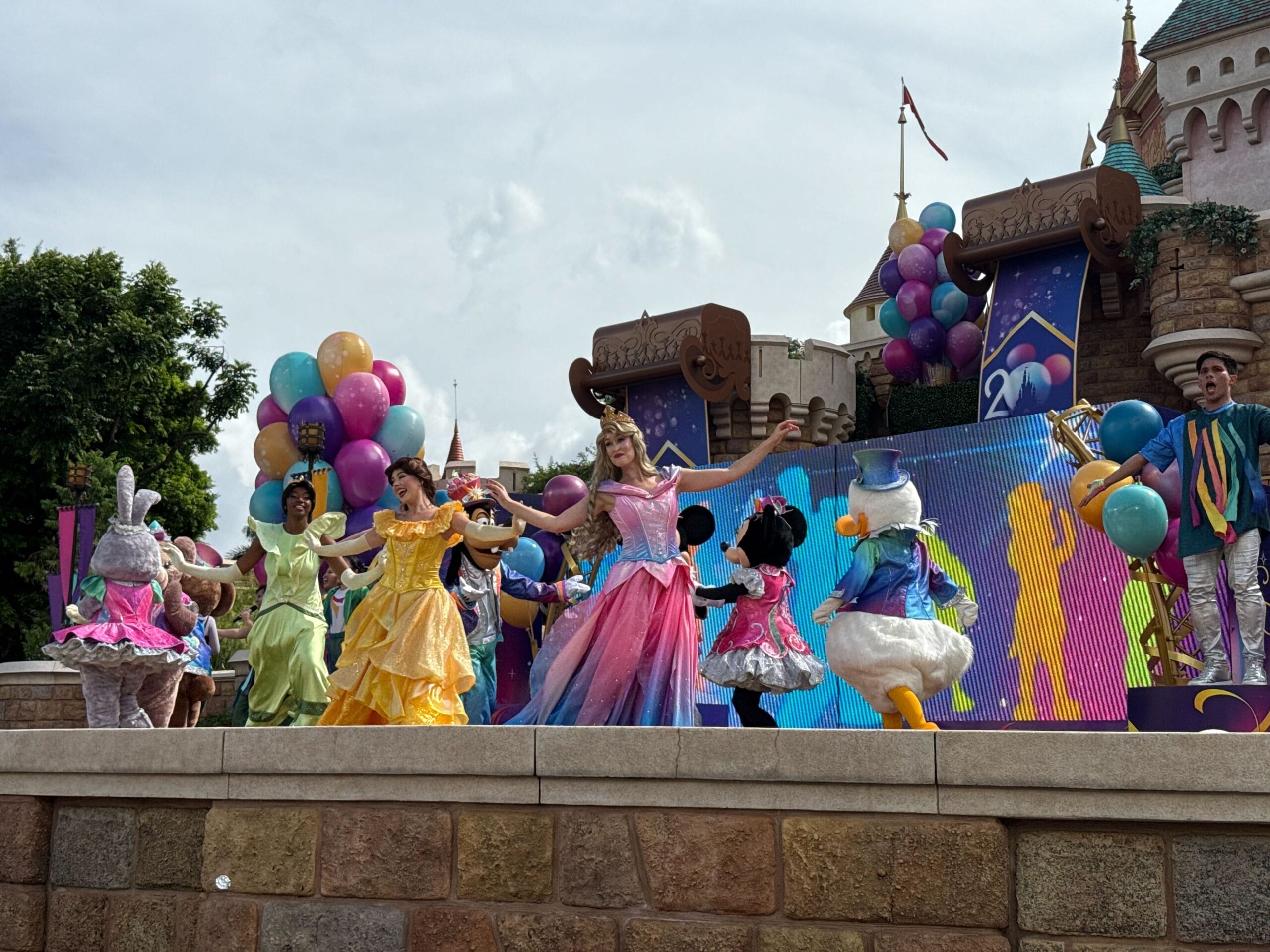 Disney characters and princesses dance on stage at the Hong Kong Disneyland 20th Anniversary Show with balloons and castle décor.