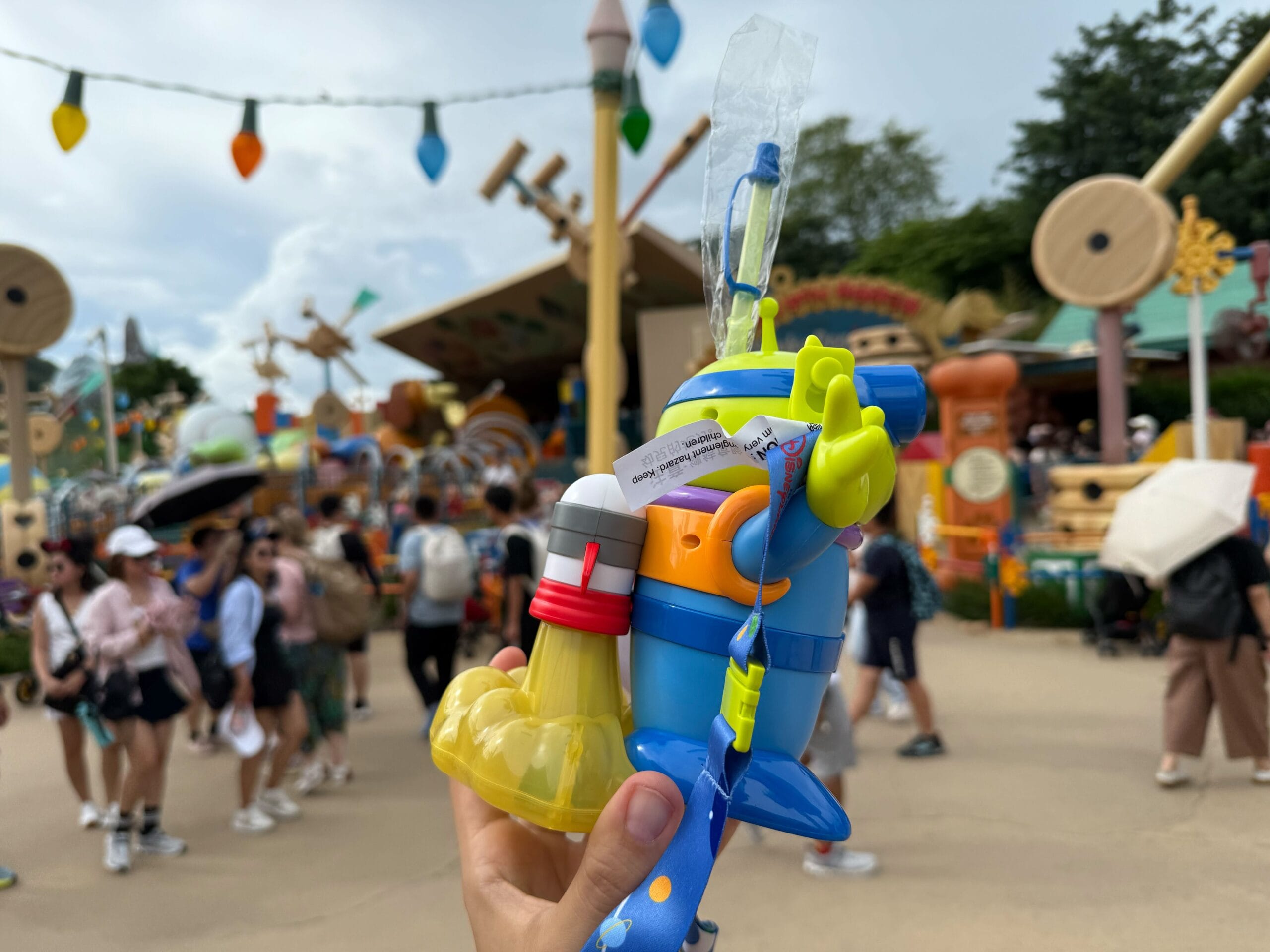 Person holding colorful toy with a straw in a busy theme park with people and decorations in the background.