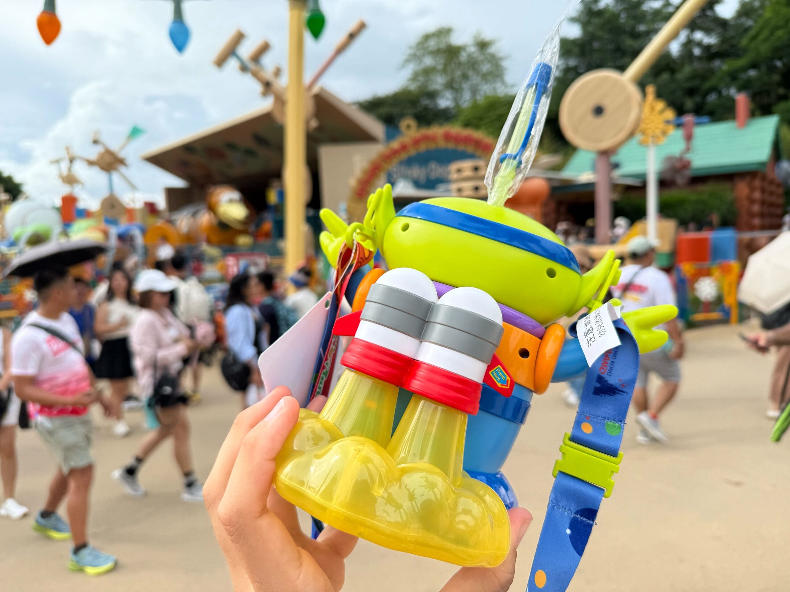 A hand holds a Toy Story alien-themed binoculars toy with people walking in an amusement park background.