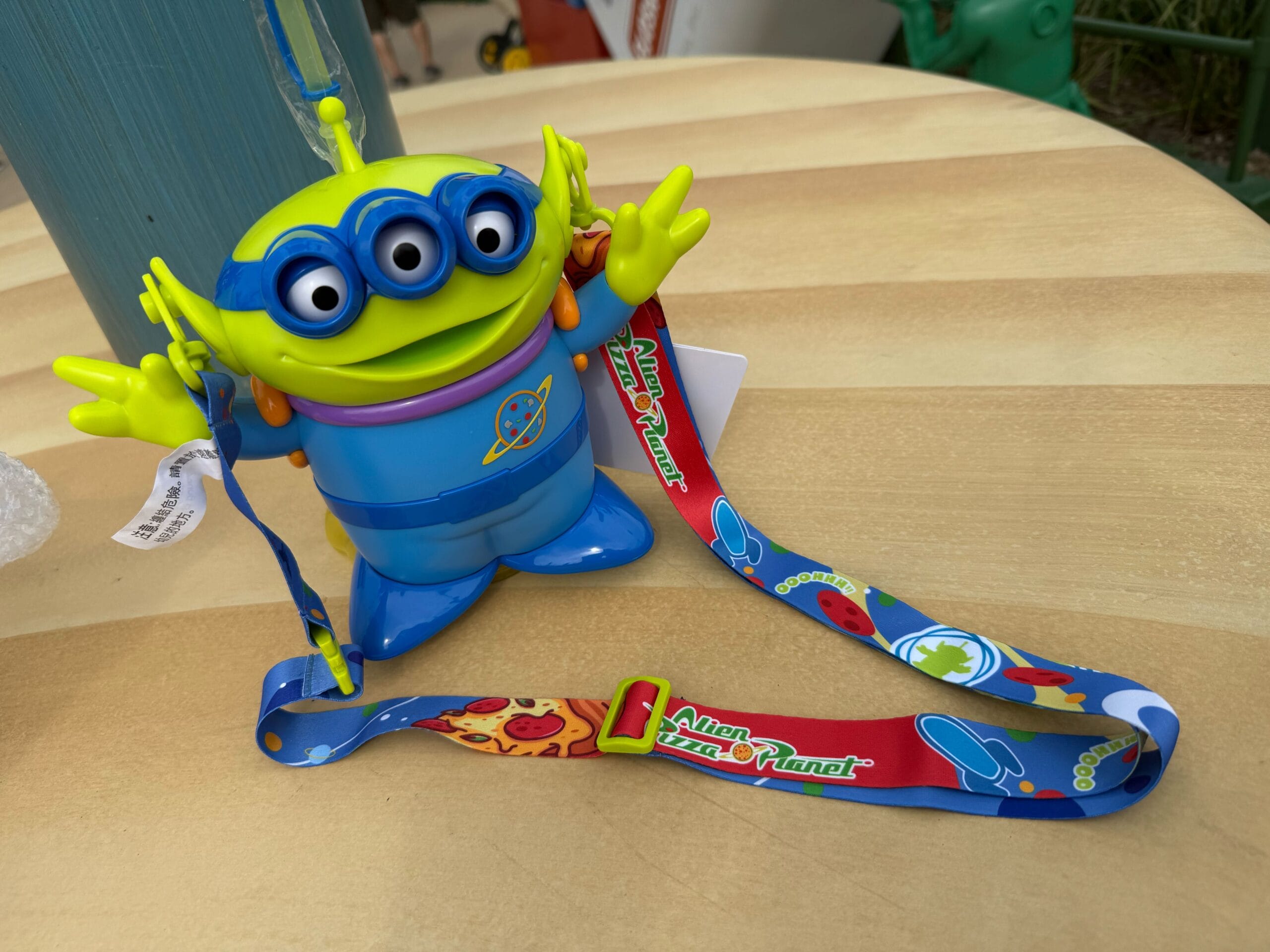 A three-eyed green alien toy with a colorful Alien Pizza Planet lanyard on a wooden table.