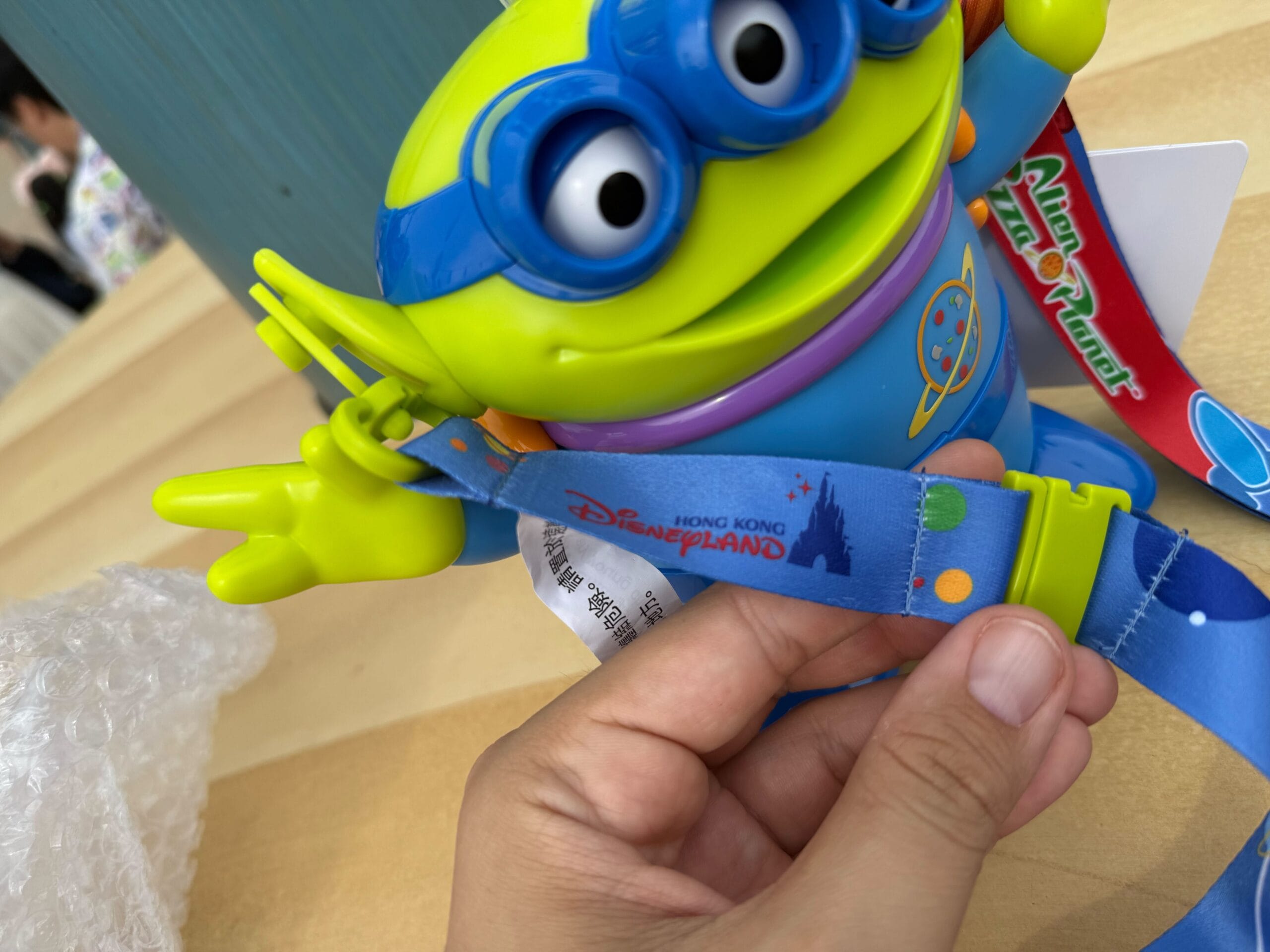 A hand holds a Hong Kong Disneyland lanyard attached to a Toy Story alien-themed souvenir cup.