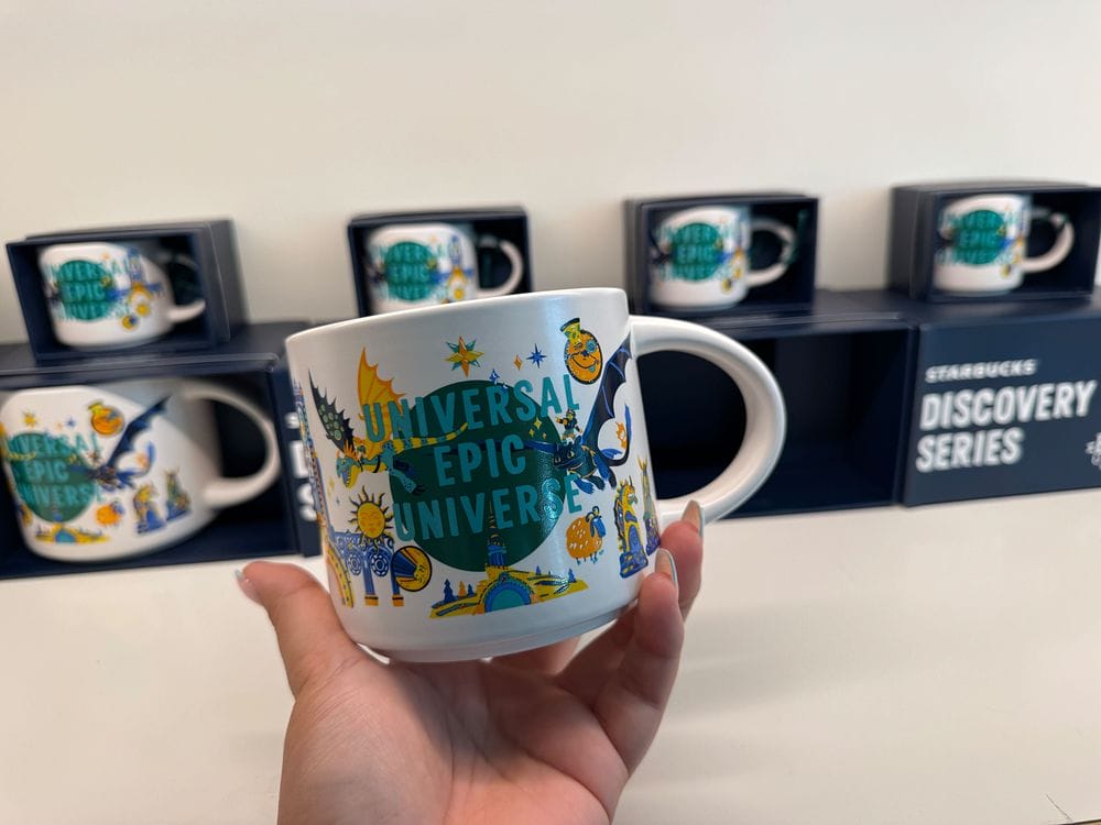 Hand holding a Starbucks "Universal Epic Universe" mug; more mugs behind. Bid in the WDWNT Summer Auction—register today!