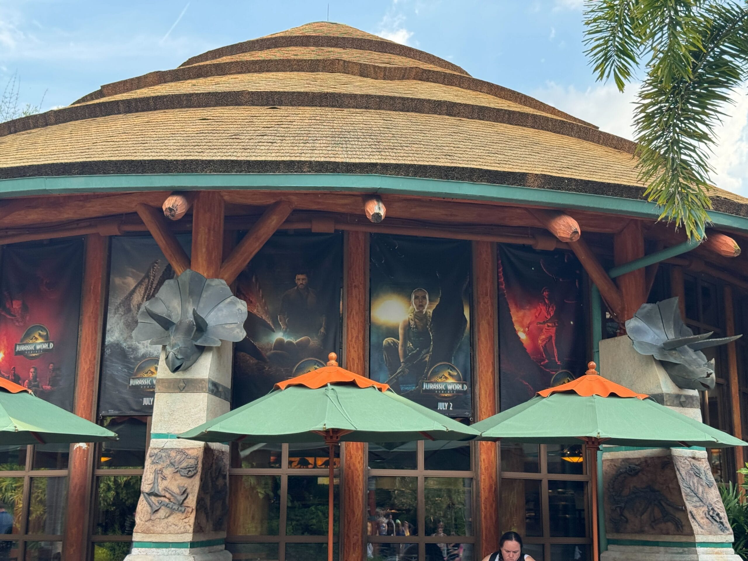 Round building with Jurassic World: Dominion movie posters and green umbrellas outside under a blue sky.