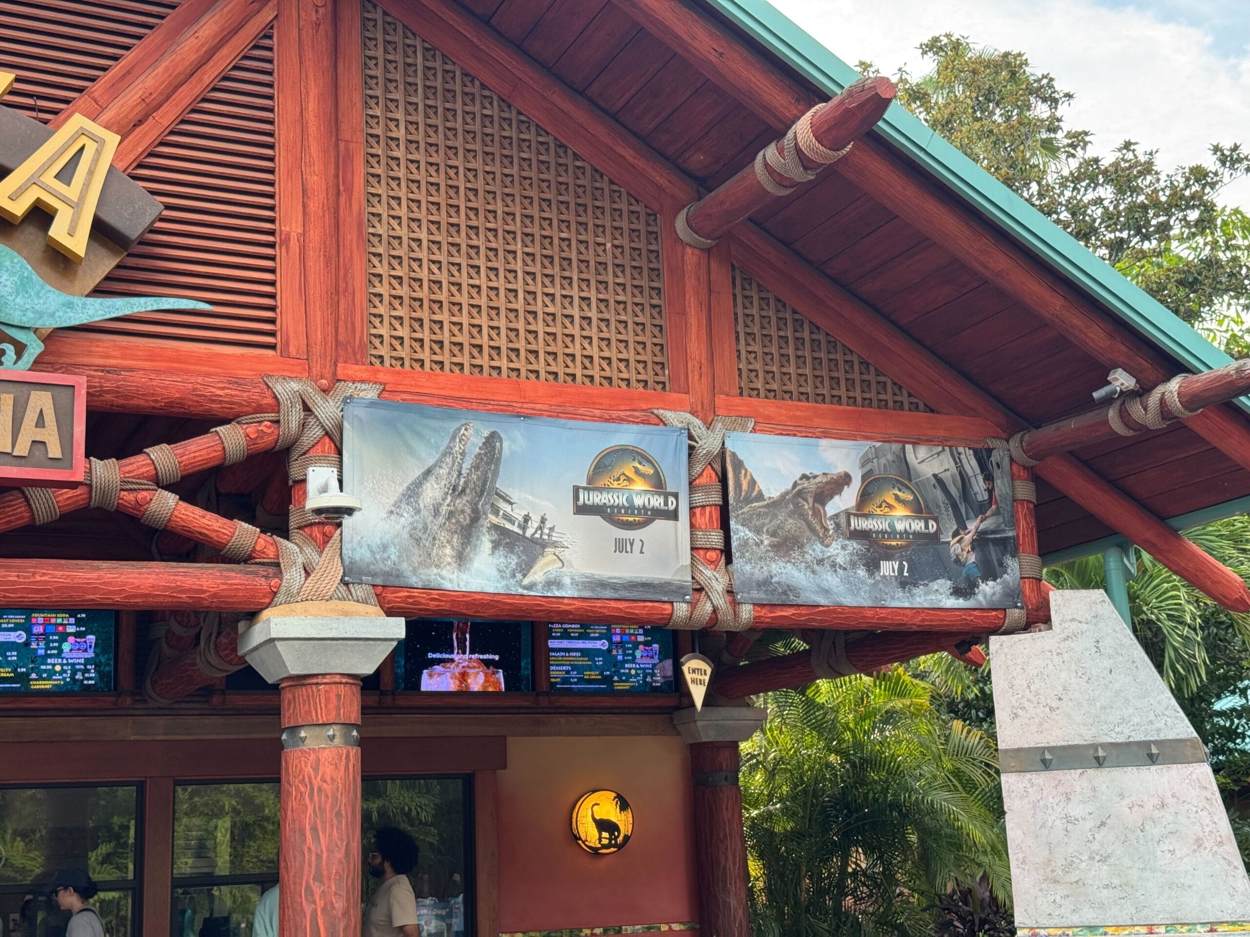 Movie posters for "Jurassic World" hang outside a wooden building with electronic screens and trees in the background.