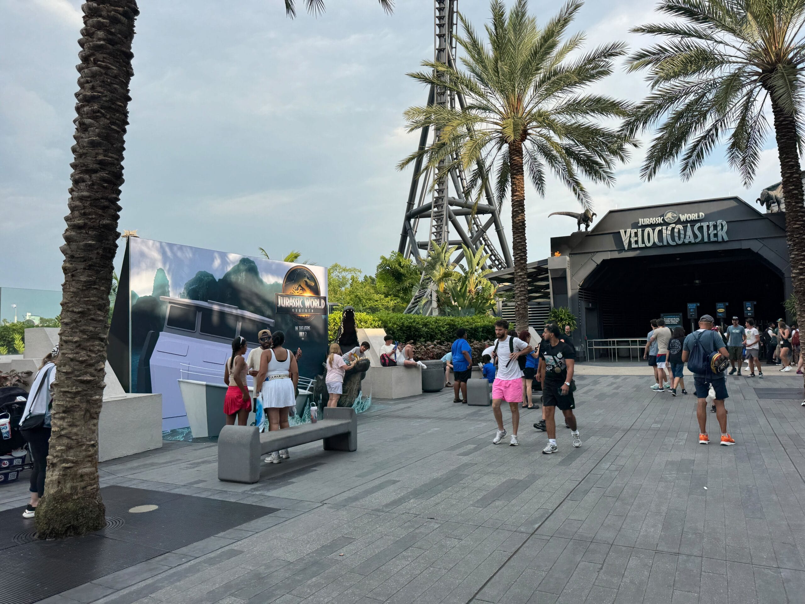 People gather near the Jurassic World VelociCoaster entrance, with palm trees and a Jurassic World sign visible.