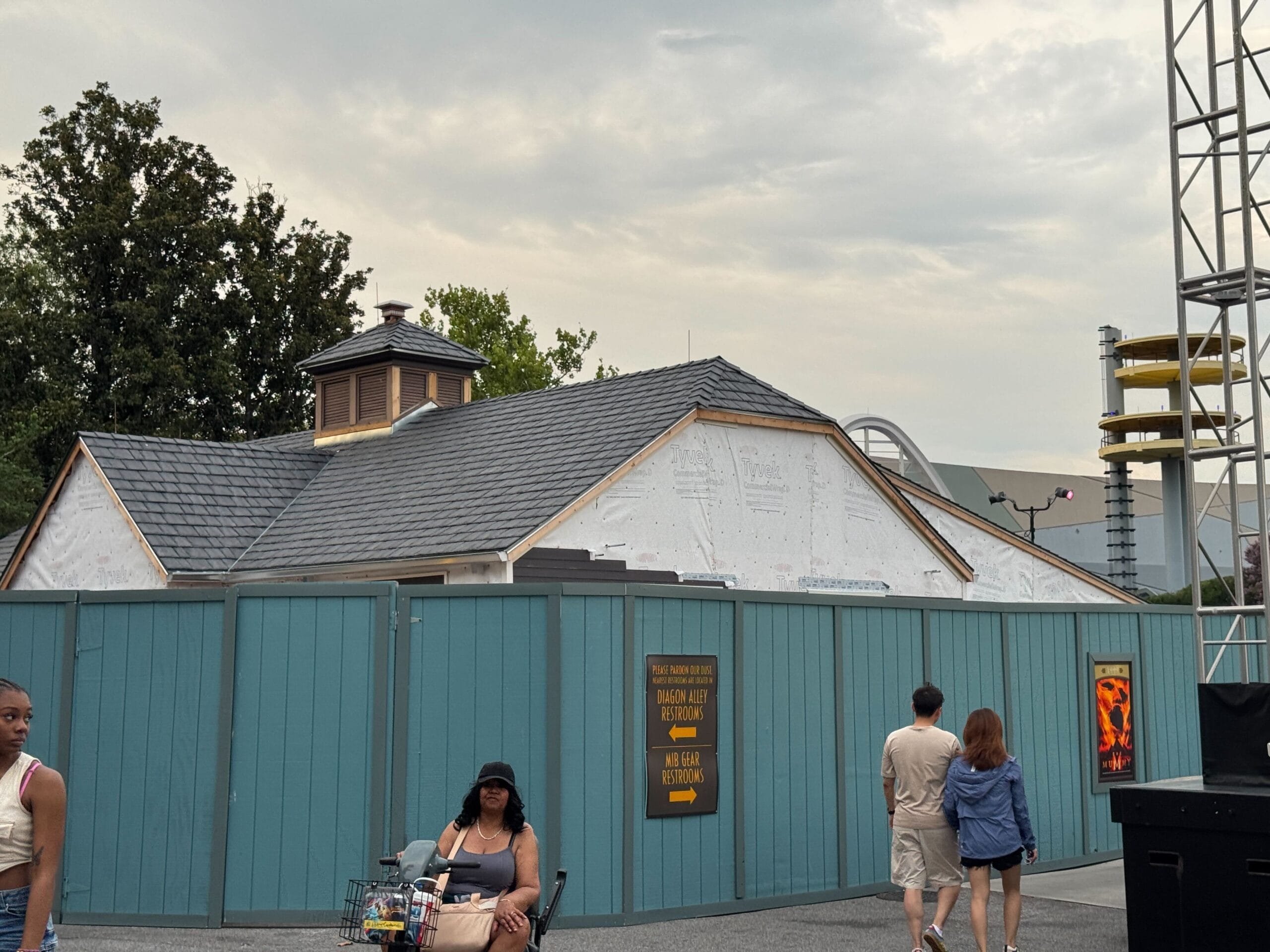 A building under construction is surrounded by blue fences; people walk by on a cloudy day.