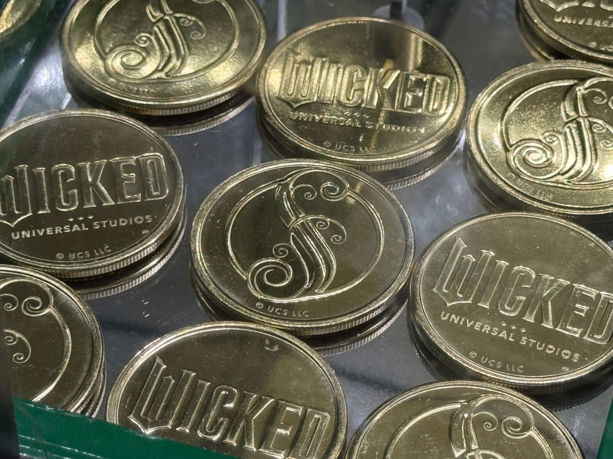 Gold coins with "Wicked" and a decorative "S" design, labeled Universal Studios, are displayed in a glass case.