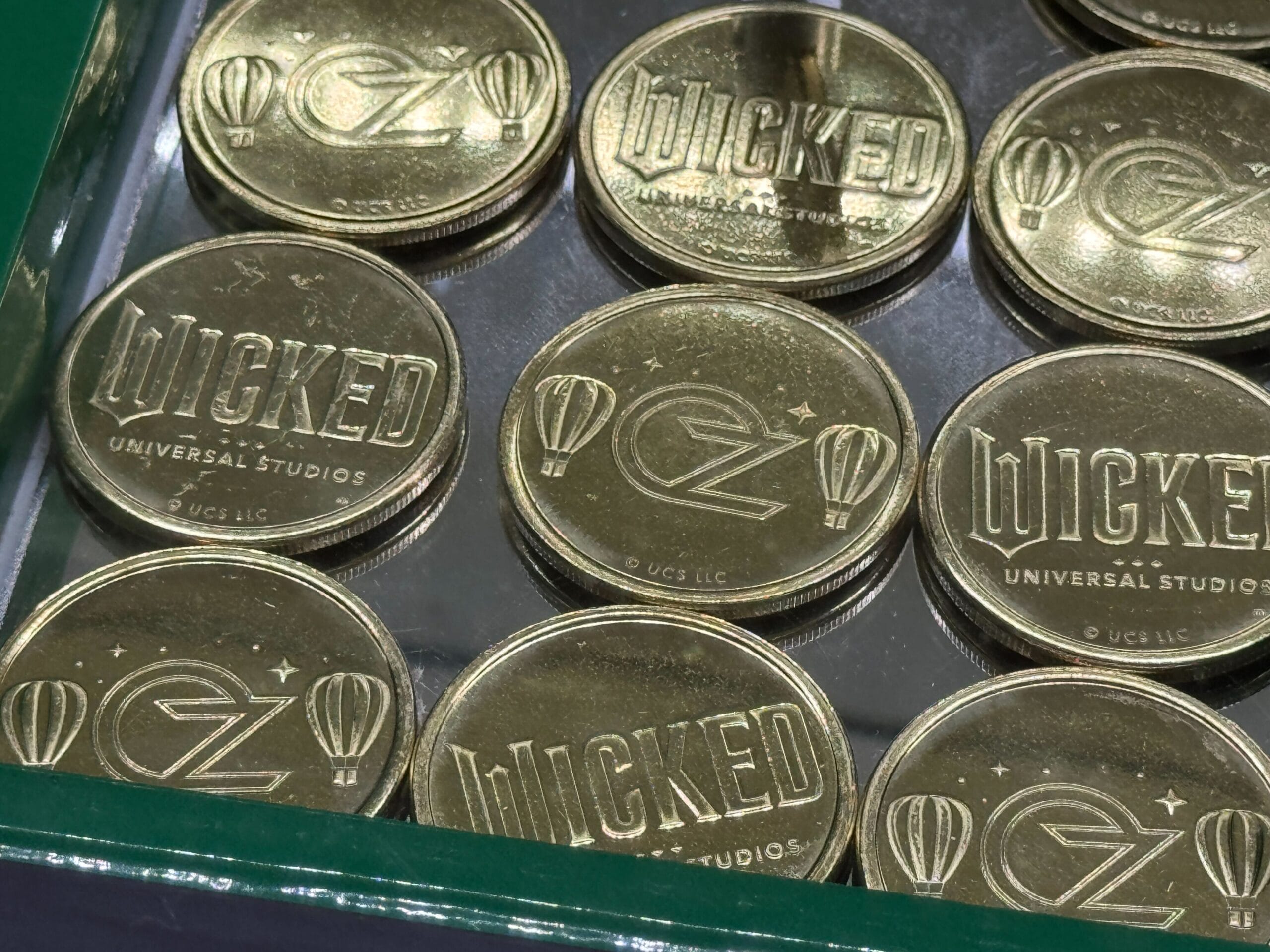 Gold coins with "WICKED" and "OZ" logos, hot air balloon symbols, and "Universal Studios" text, stacked in a display case.