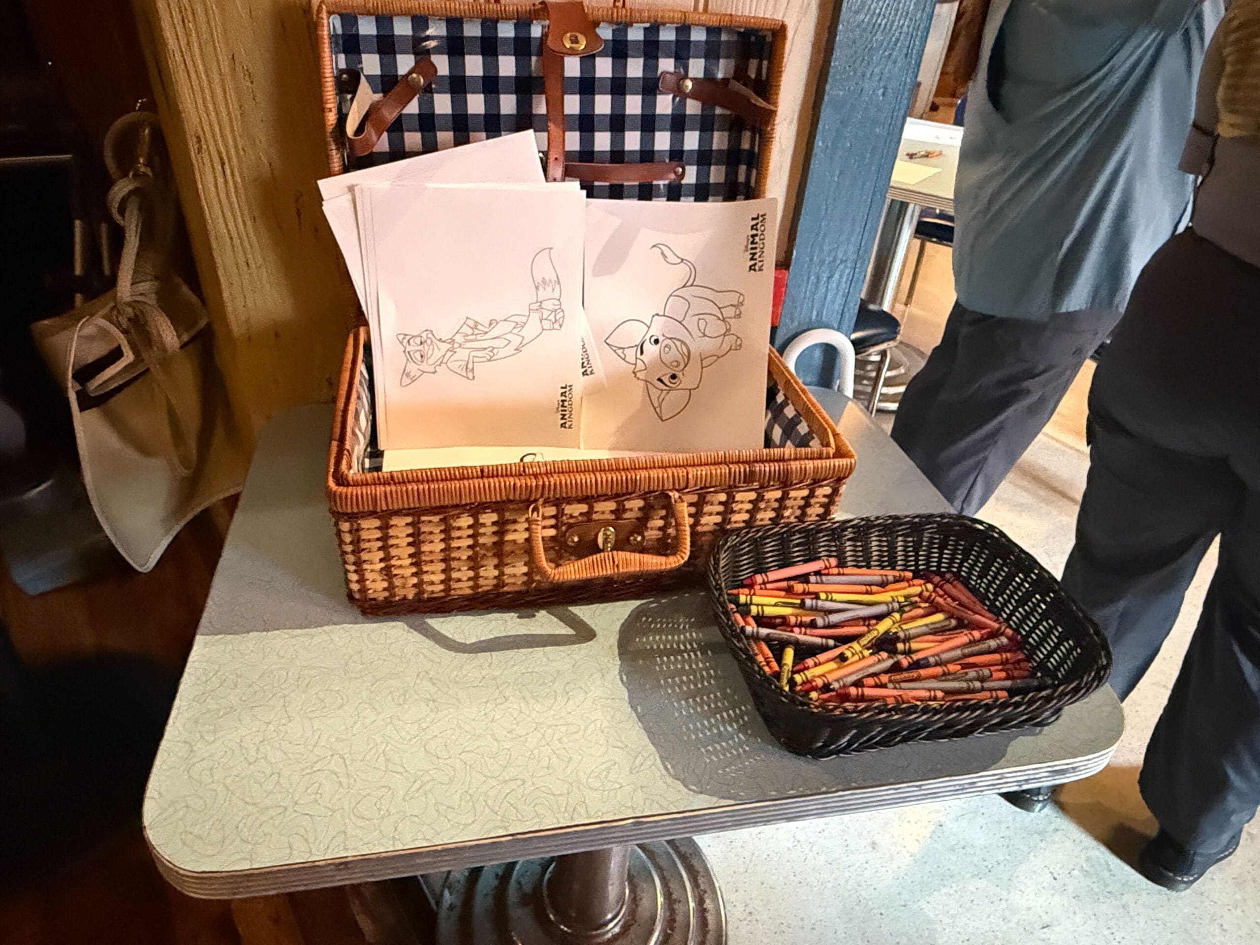 A basket with coloring pages and a smaller basket of crayons sits on a table in a cozy indoor setting.