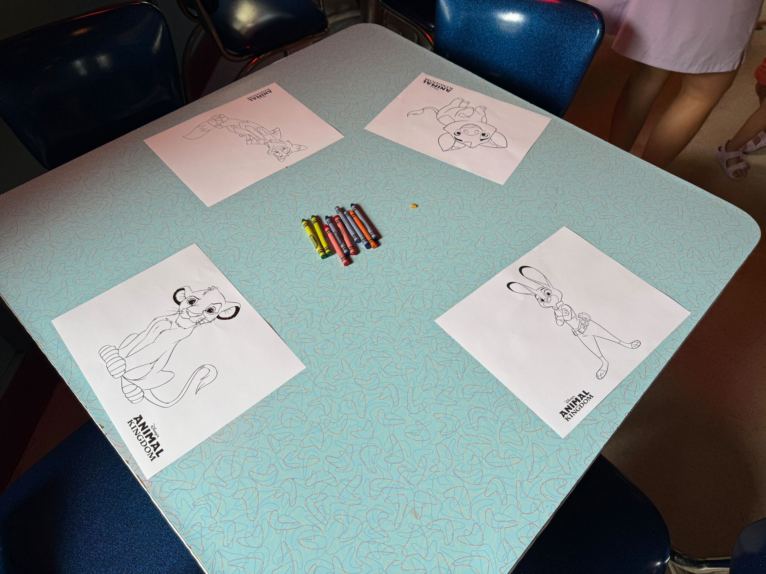 A table with crayons and four animal coloring pages, each with a different Disney character outlined in black.