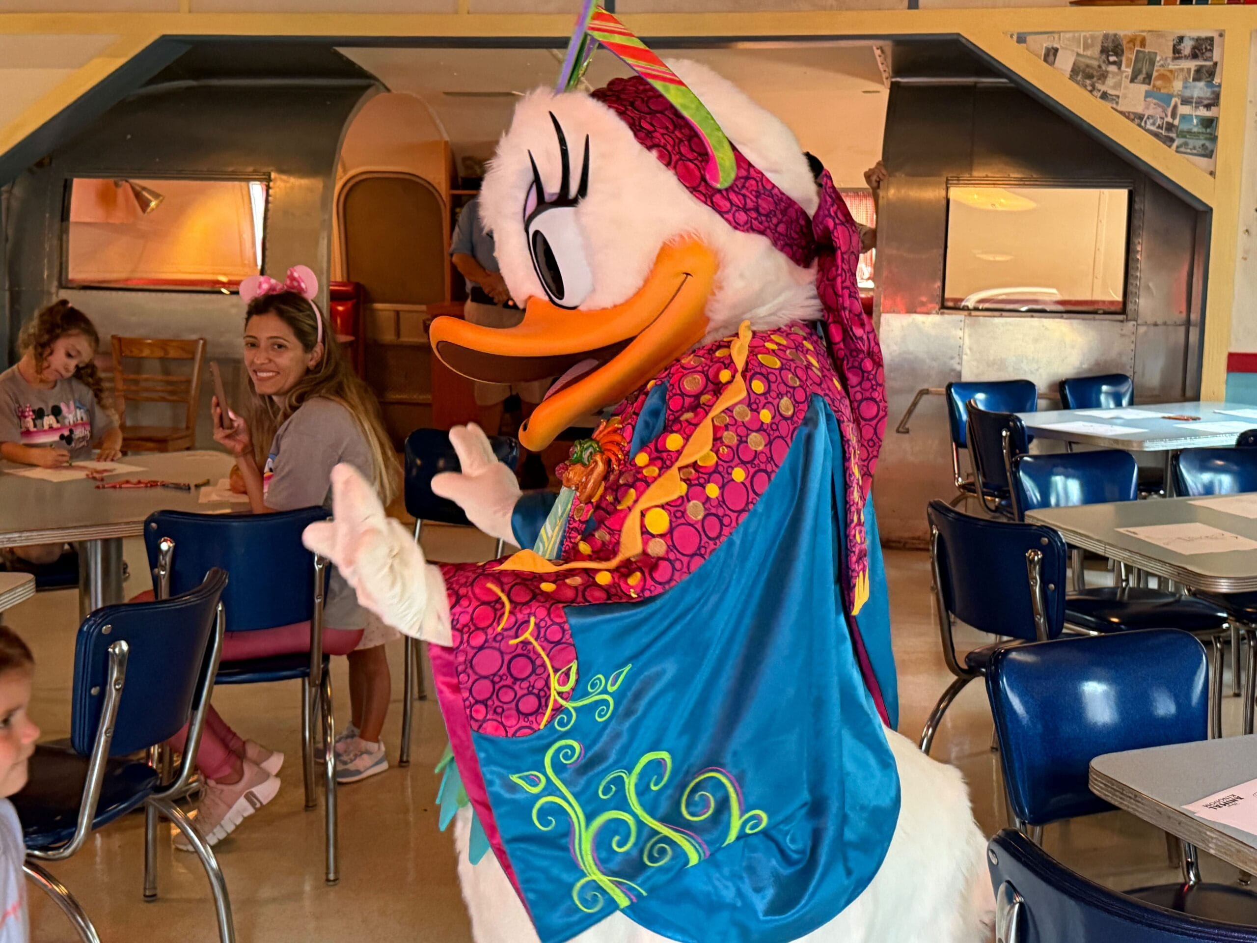 Daisy Duck in a colorful outfit greets children in a retro-themed restaurant, while guests smile in the background.