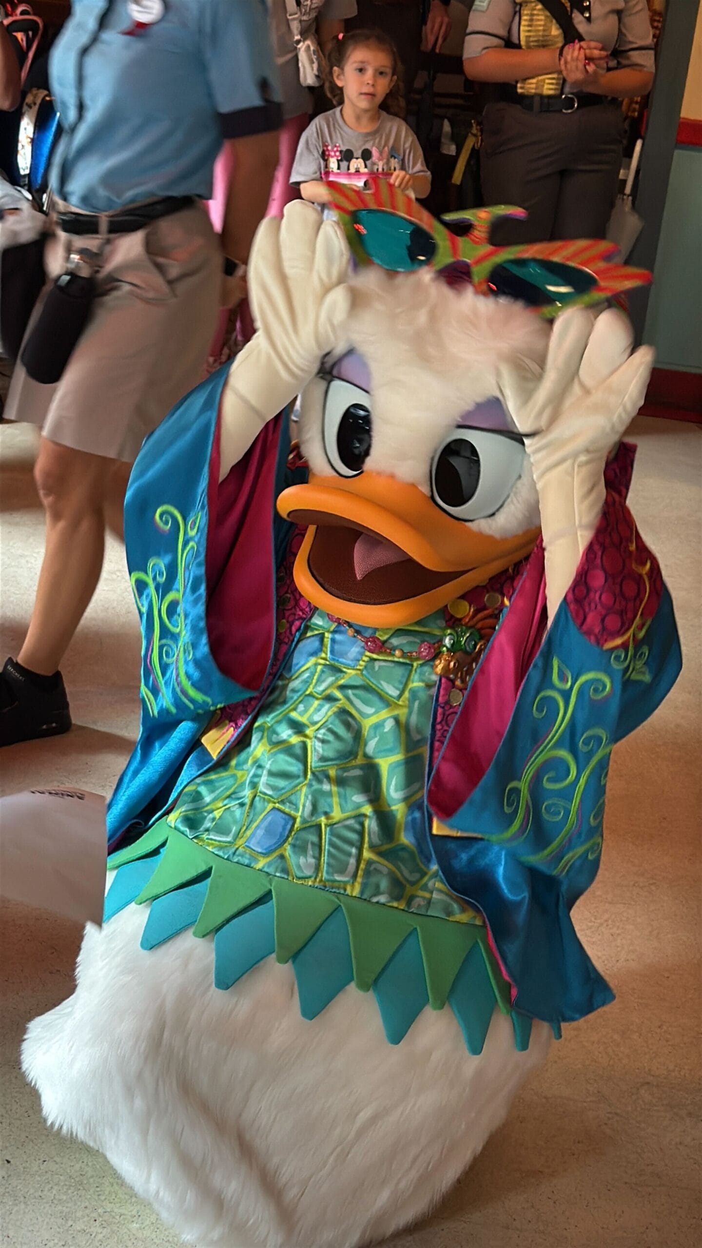 Daisy Duck in a colorful costume poses for photos at Daisy's Dig, surrounded by people at an indoor event.