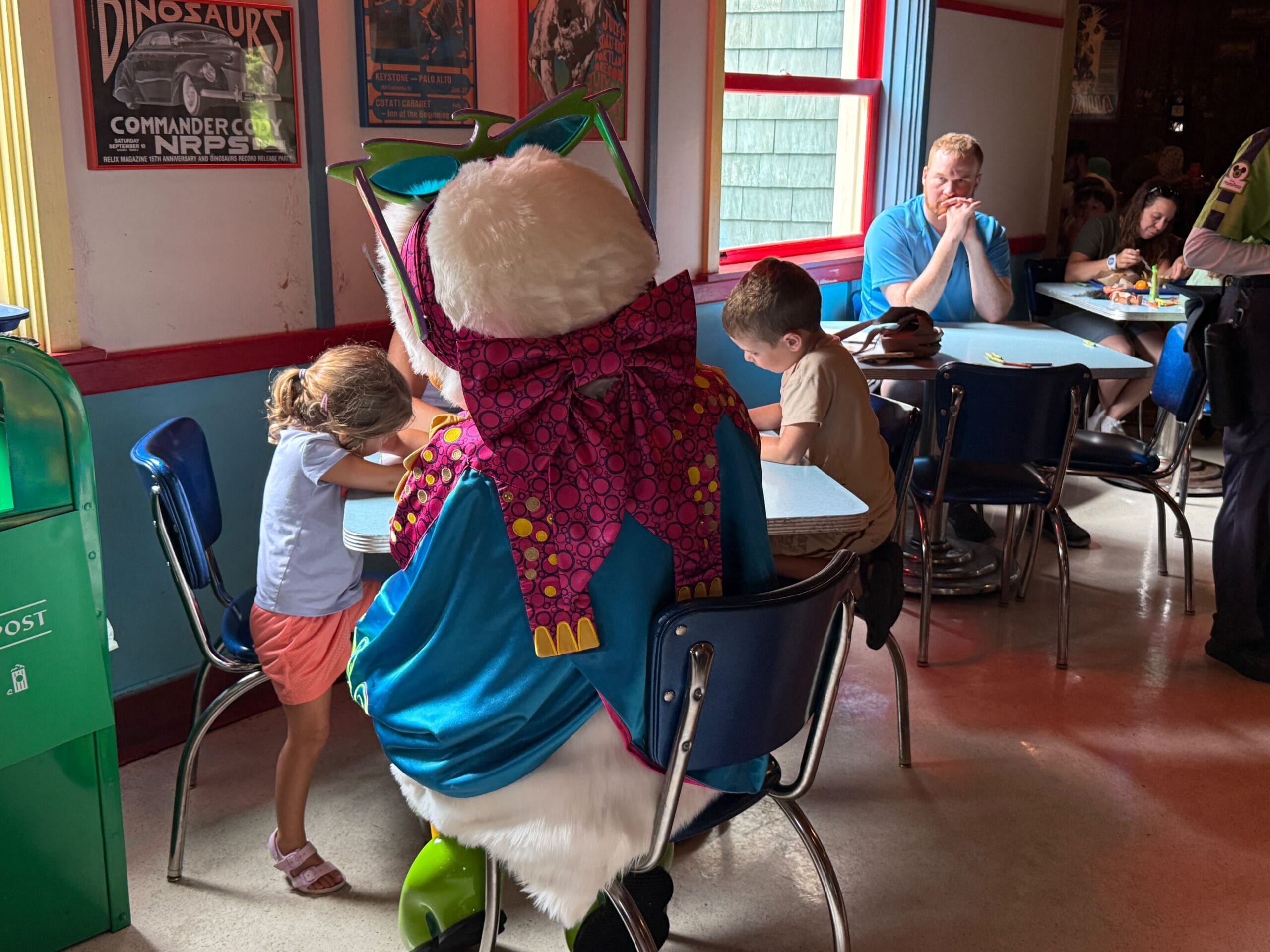 A person in a bunny costume sits with two children coloring at Daisy's Dig in a brightly lit restaurant.