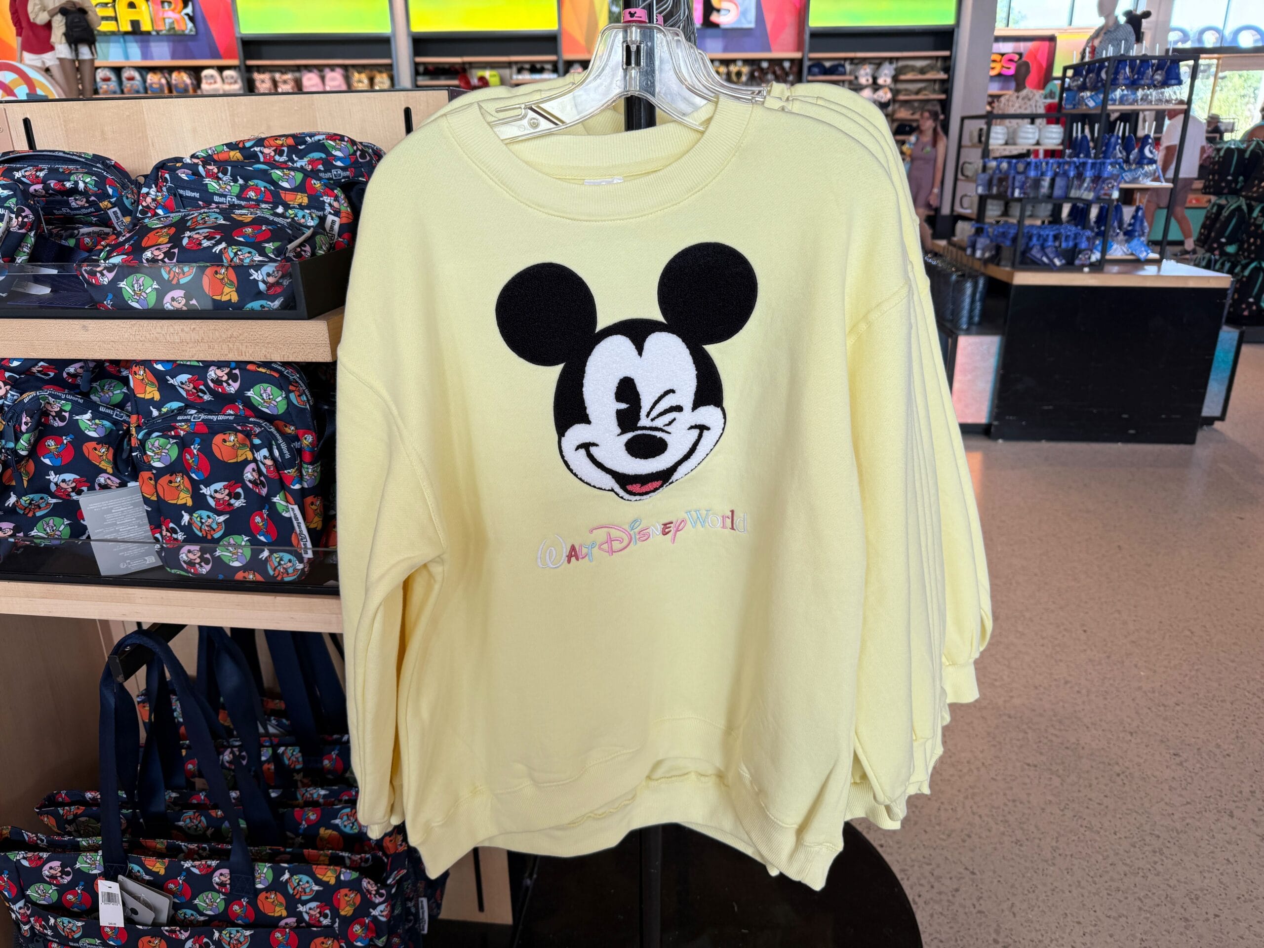 A yellow Character Sweatshirt with a winking Mickey and "Walt Disney World" text hangs on a store rack.
