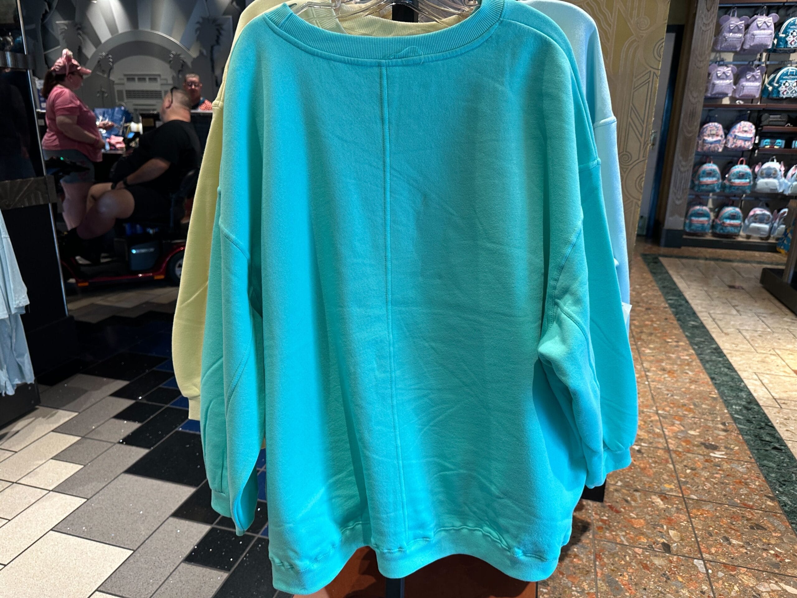 A turquoise Walt Disney World Character Sweatshirt is displayed on a rack in a retail store with shoppers in the background.