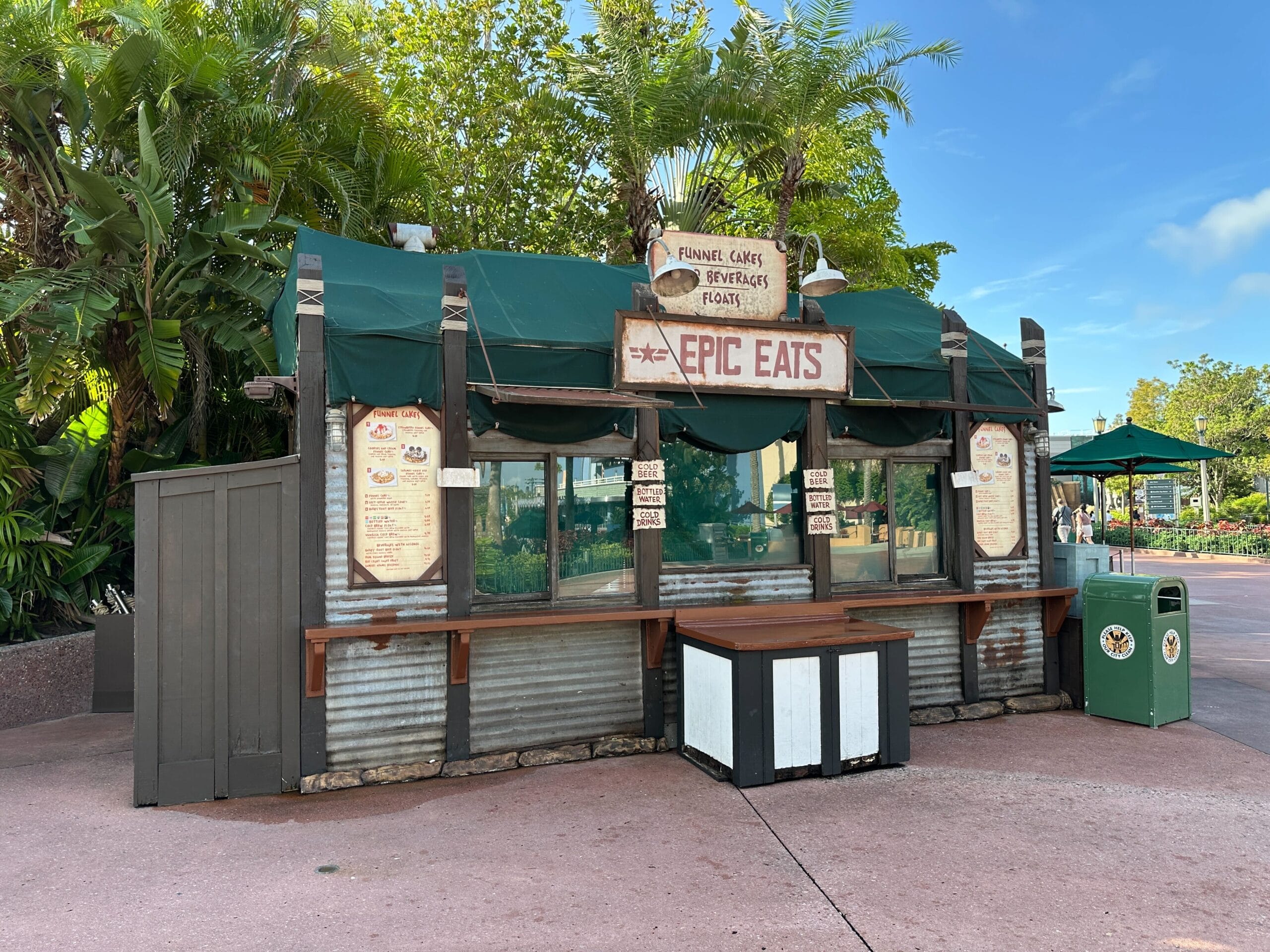 A closed outdoor food stand named "Epic Eats" with a green awning and menu signs, surrounded by palm trees.