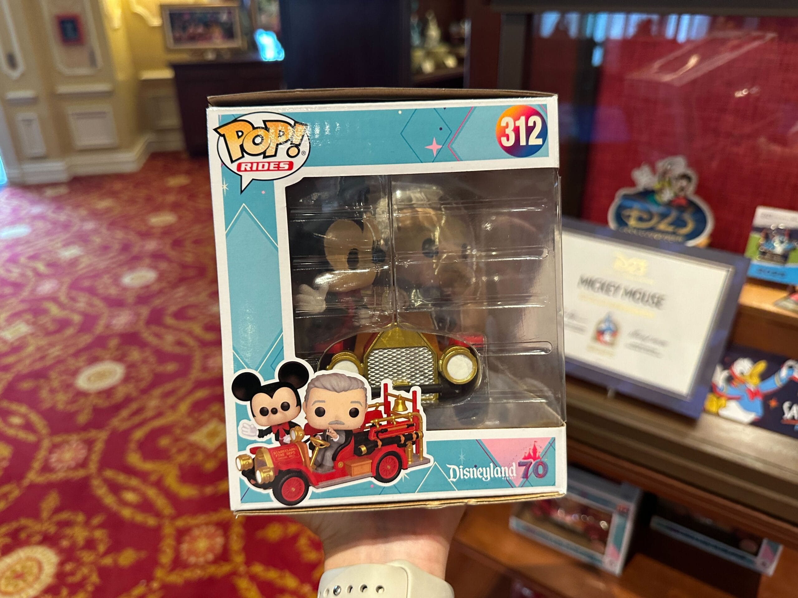 A hand holds a Pop! Rides figure of Mickey Mouse in a red car, labeled "Disneyland 70th," inside a store.