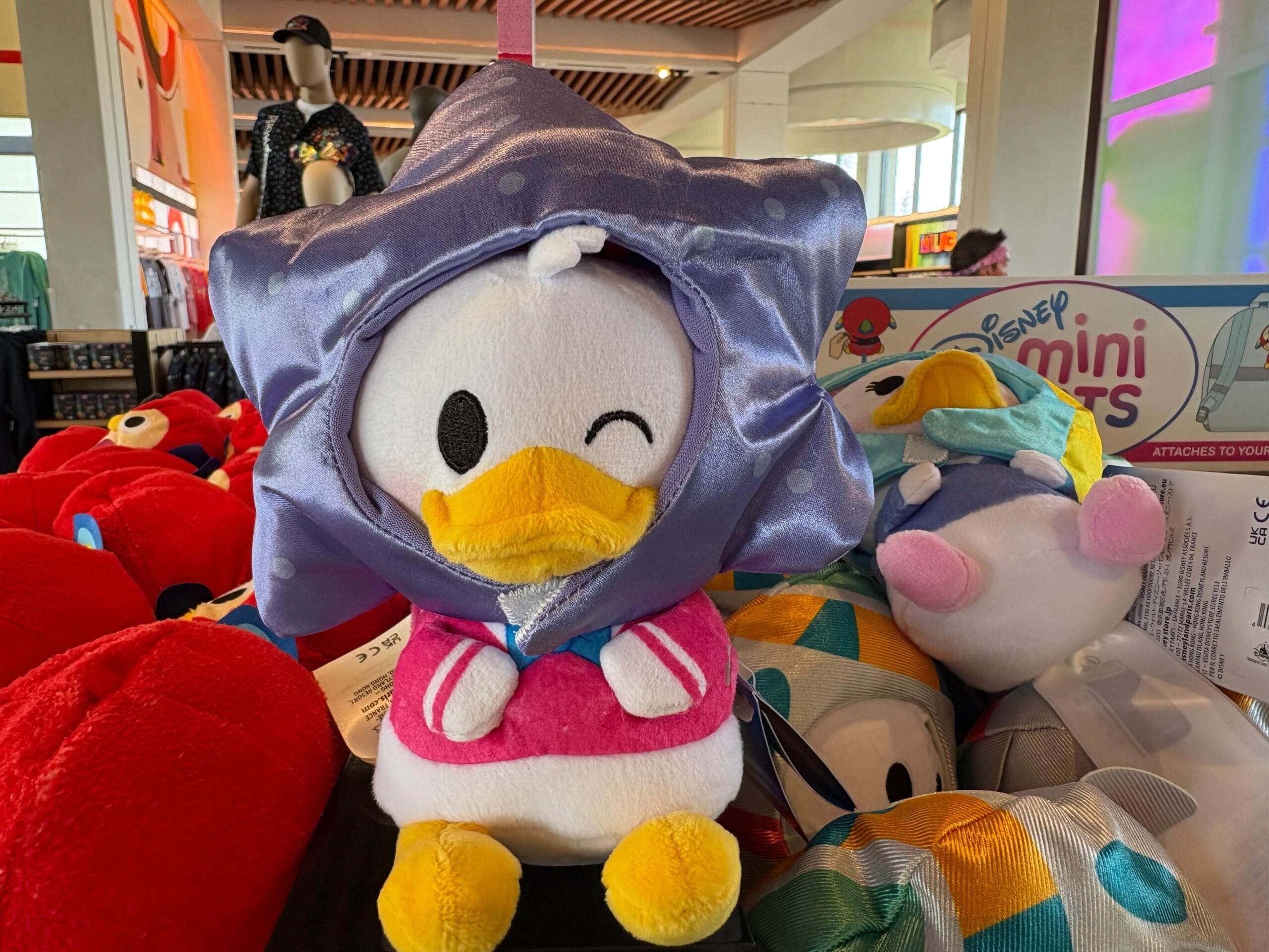Plush toy of Daisy Duck winking, wearing a purple star-shaped hood, displayed among other colorful stuffed toys.