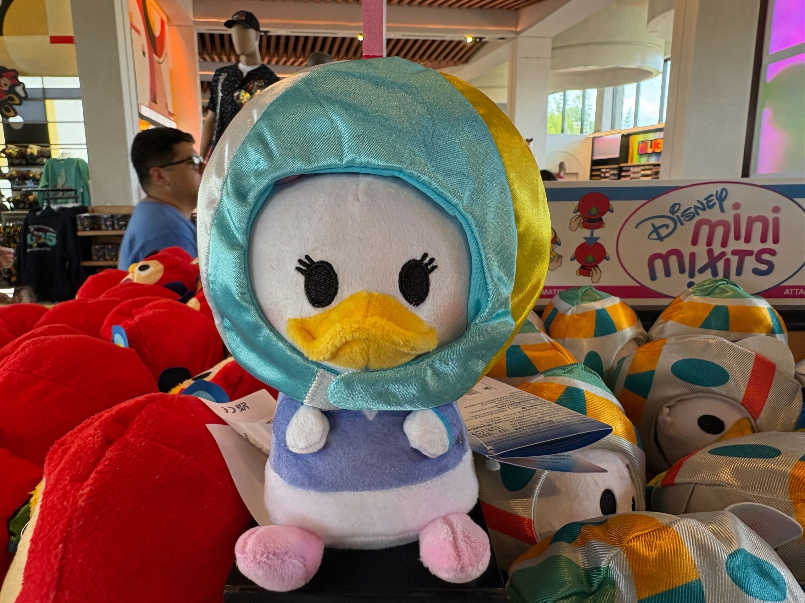 A plush toy of Daisy Duck in a blue hood on display among other Disney Mini Maits plushies in a store.