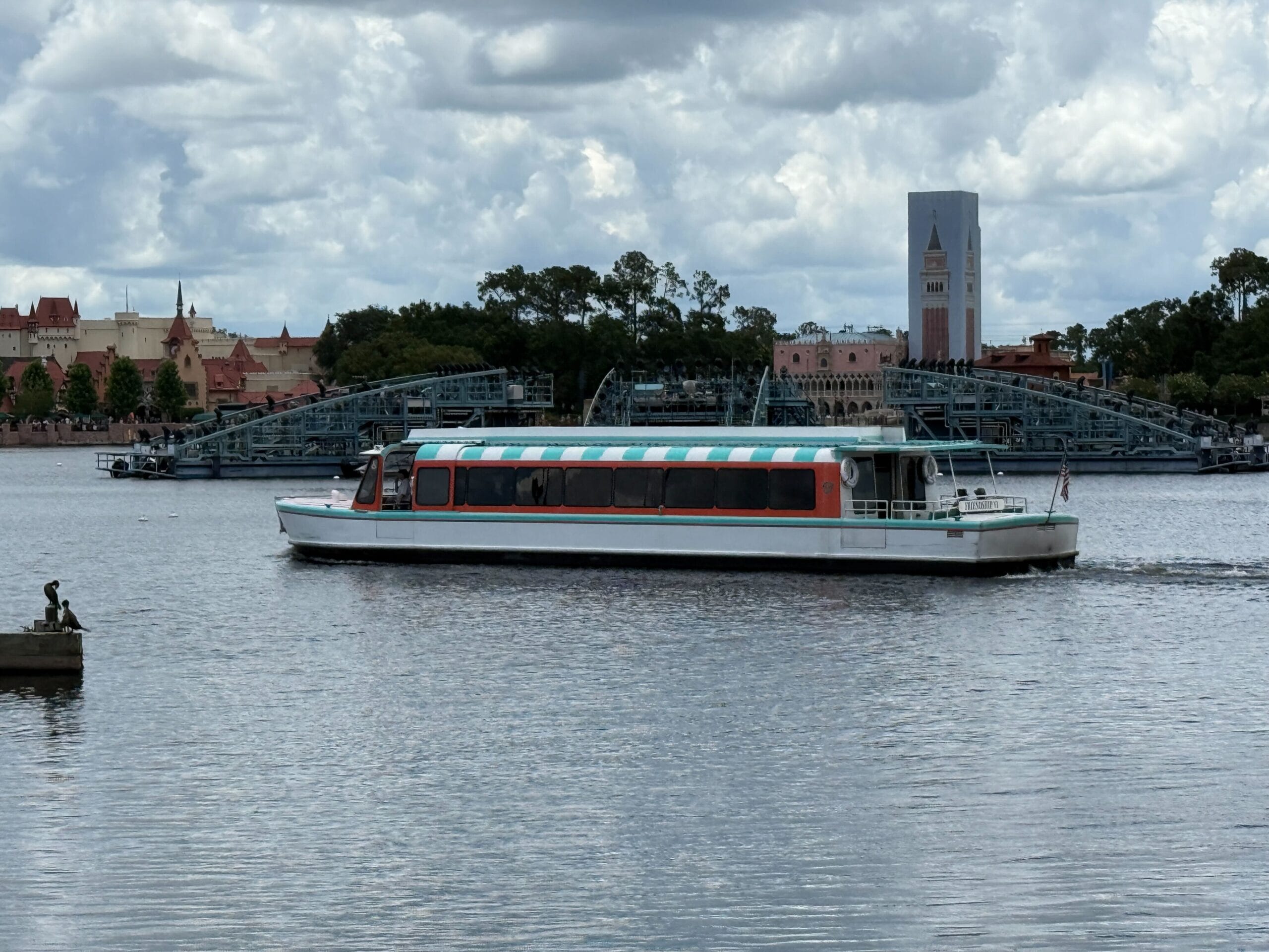 A white and orange Friendship Boat crosses the lake at EPCOT’s World Showcase with buildings and cloudy sky behind.
