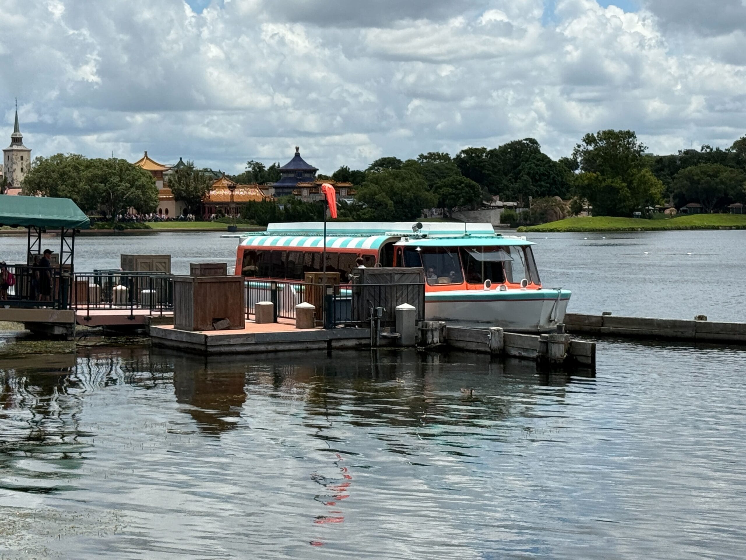 A retro-style Friendship Boat with teal and orange accents is docked on a calm lake at EPCOT’s World Showcase.