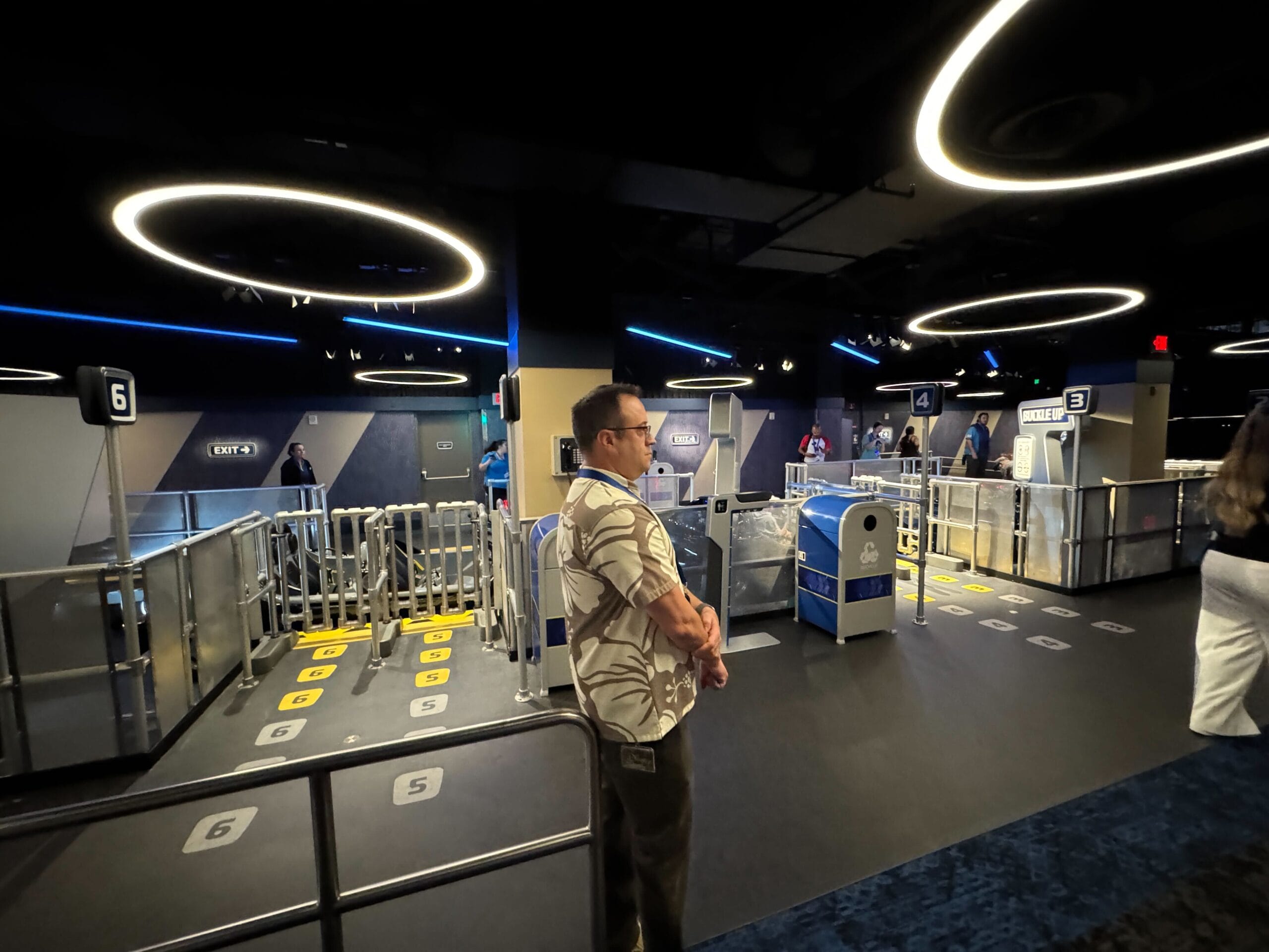 A man stands in a futuristic, neon-lit Test Track queue area with numbered gates and circular ceiling lights.
