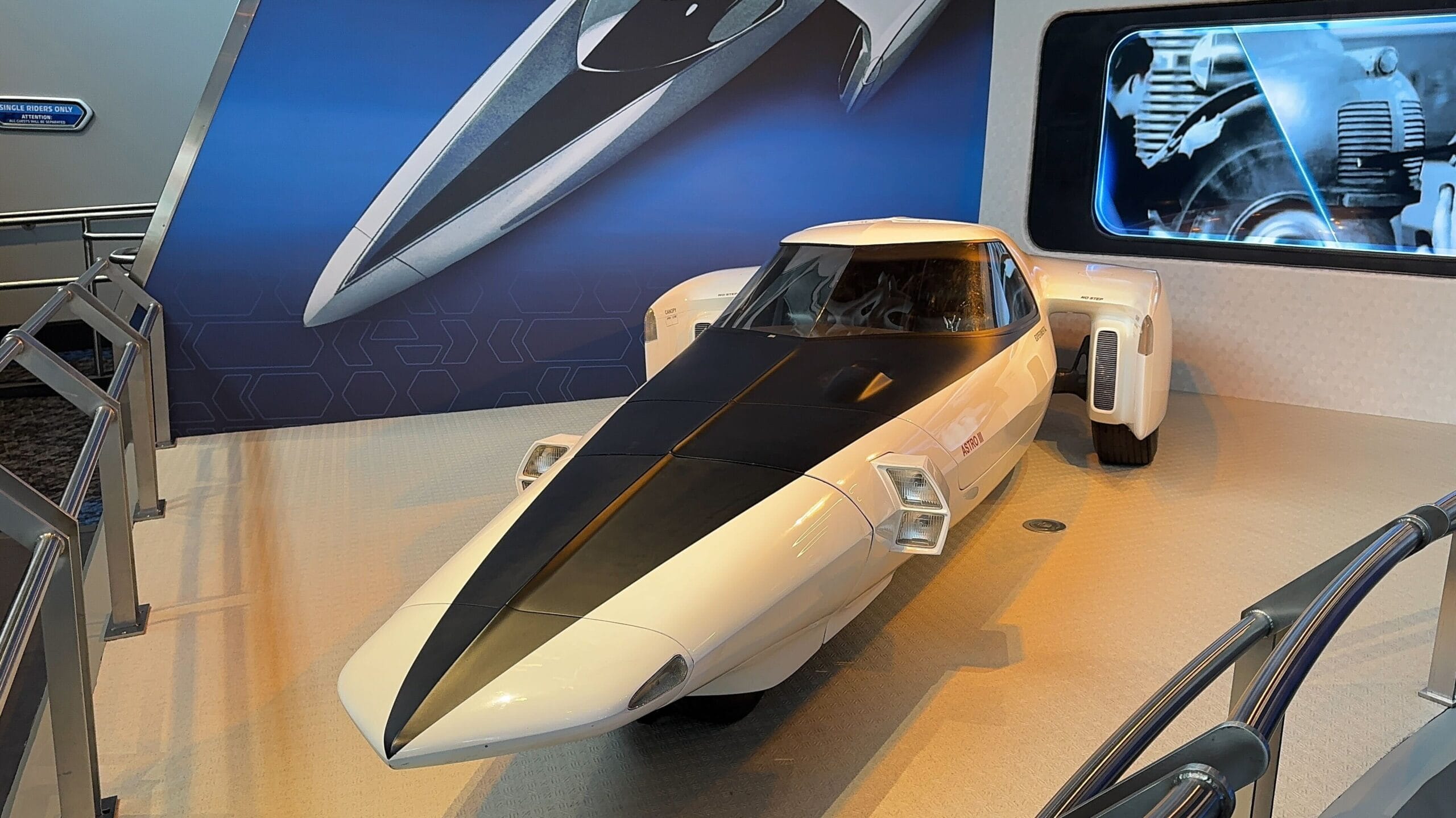 Futuristic white and black car on display indoors with PHOTOS and a matching car image on the wall behind it.