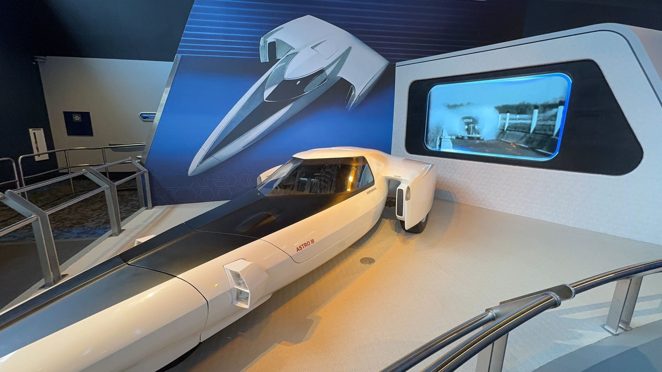 White futuristic car display with Test Track concept illustration and a screen showing a dynamic driving scene in the background.