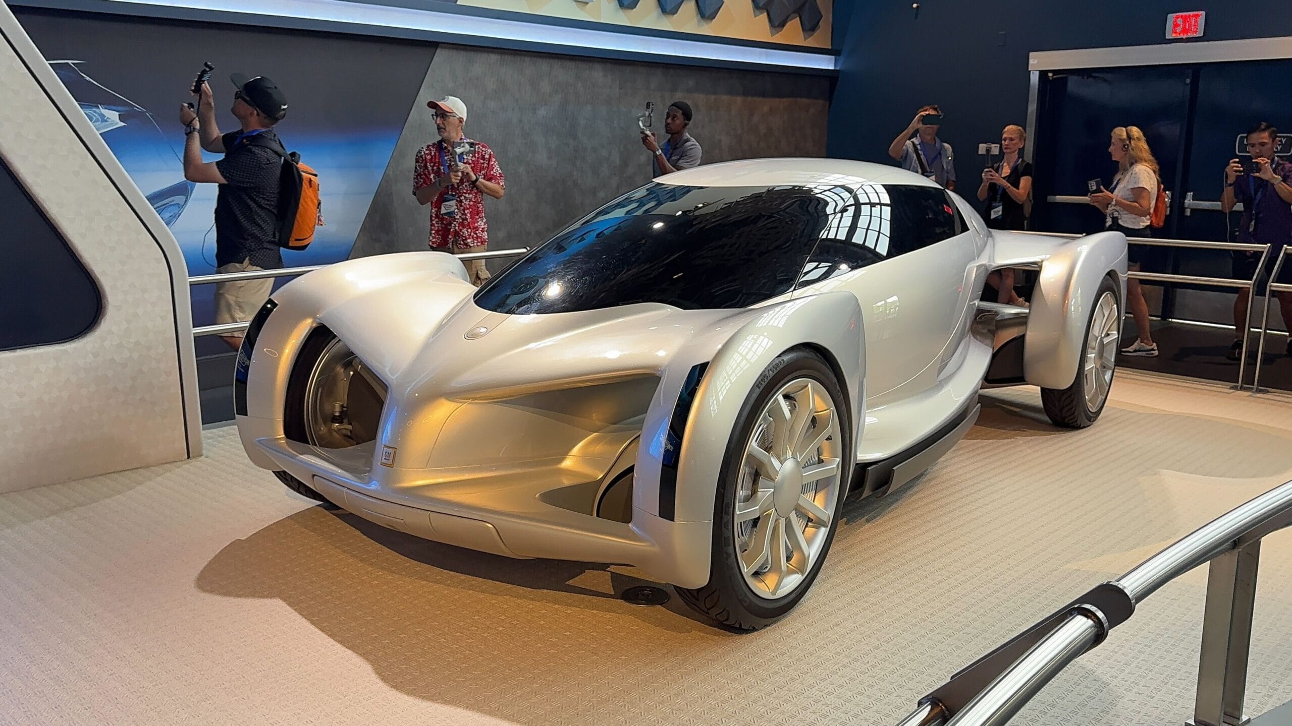 A futuristic silver concept car on display indoors at Test Track, with people taking photos around it.