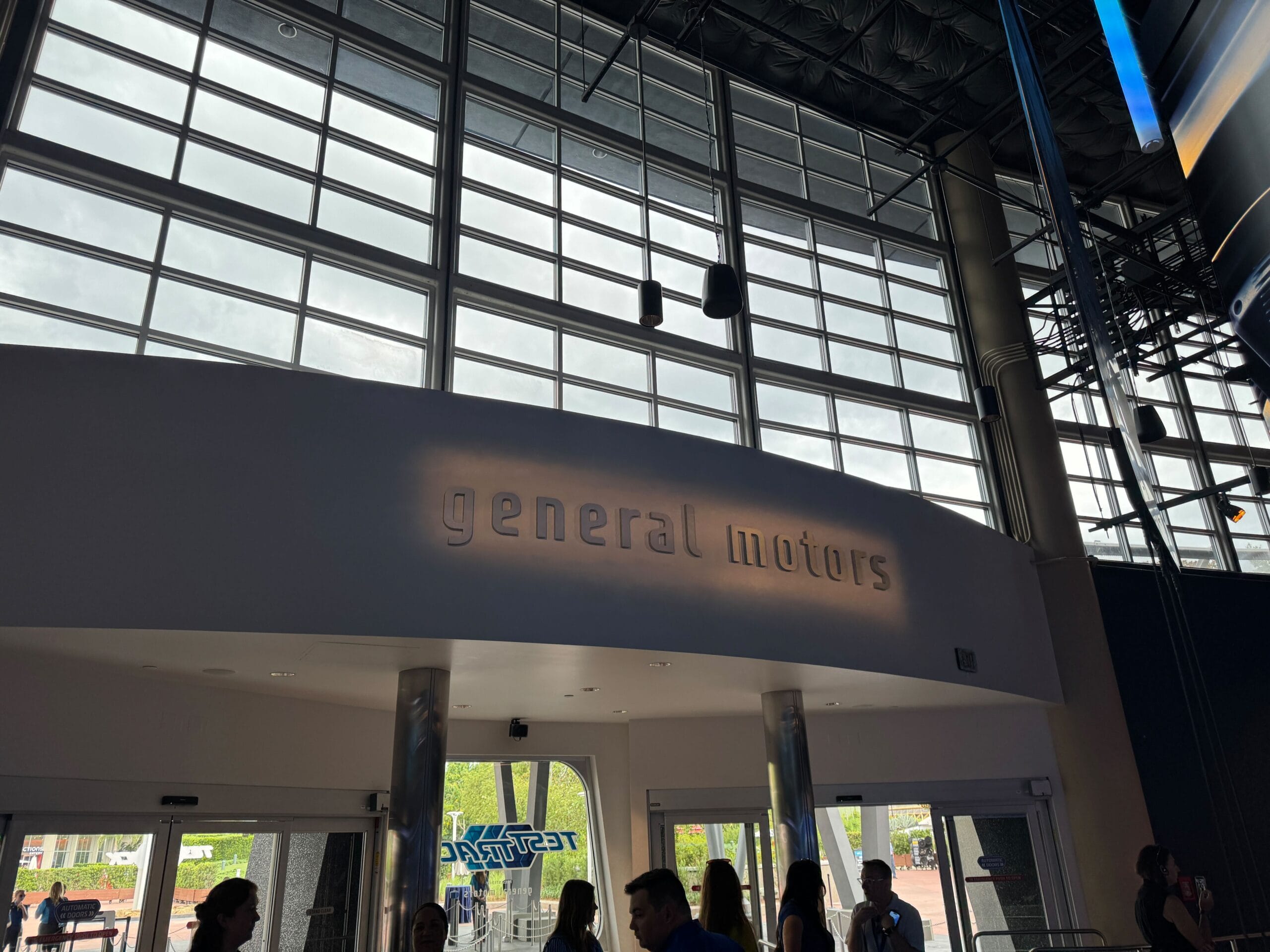 Silhouetted people stand beneath a "general motors" sign near the Test Track inside a modern building with large glass windows.