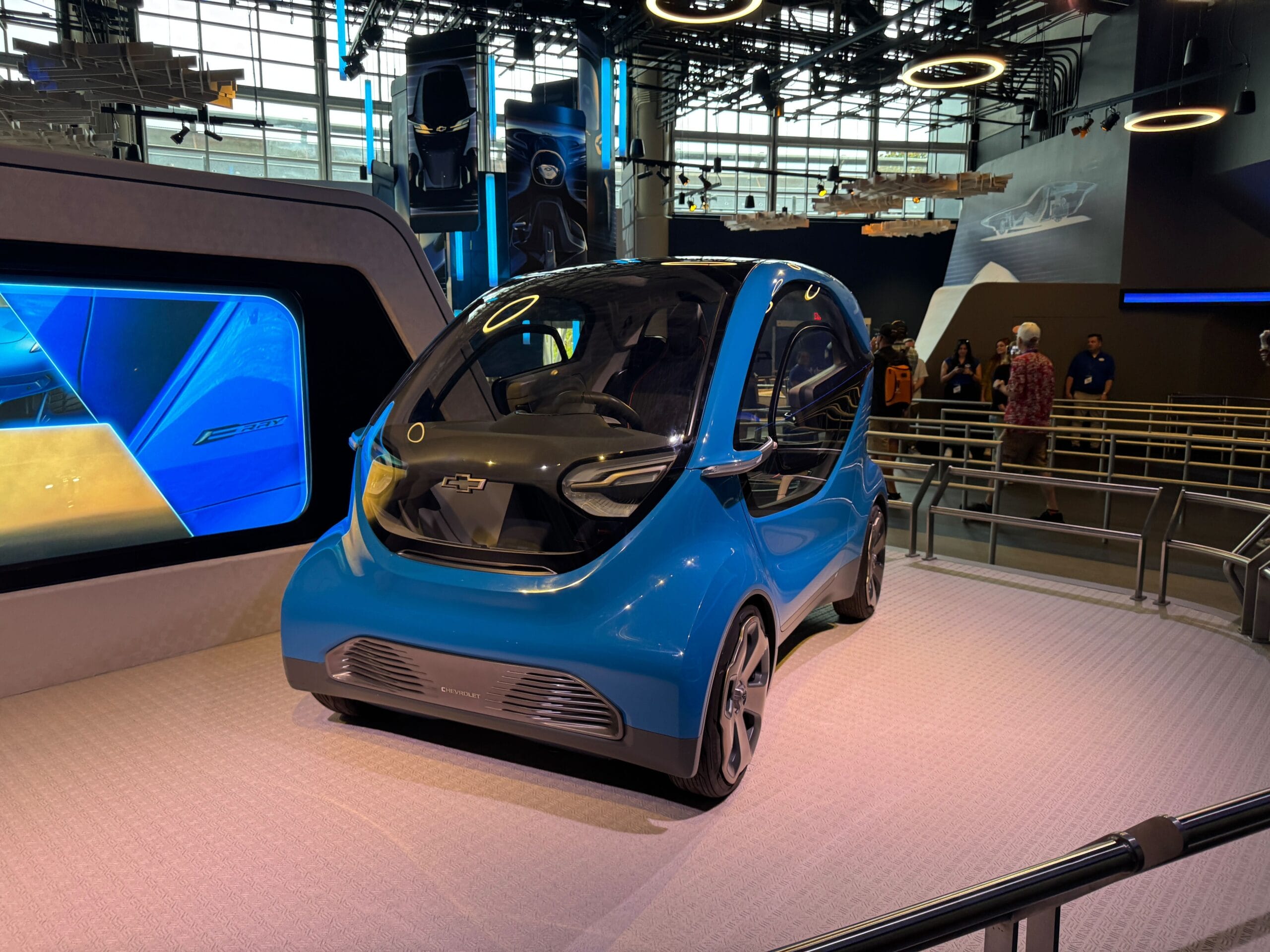 A small, blue, futuristic Test Track concept car is displayed indoors with people and digital screens in the background.
