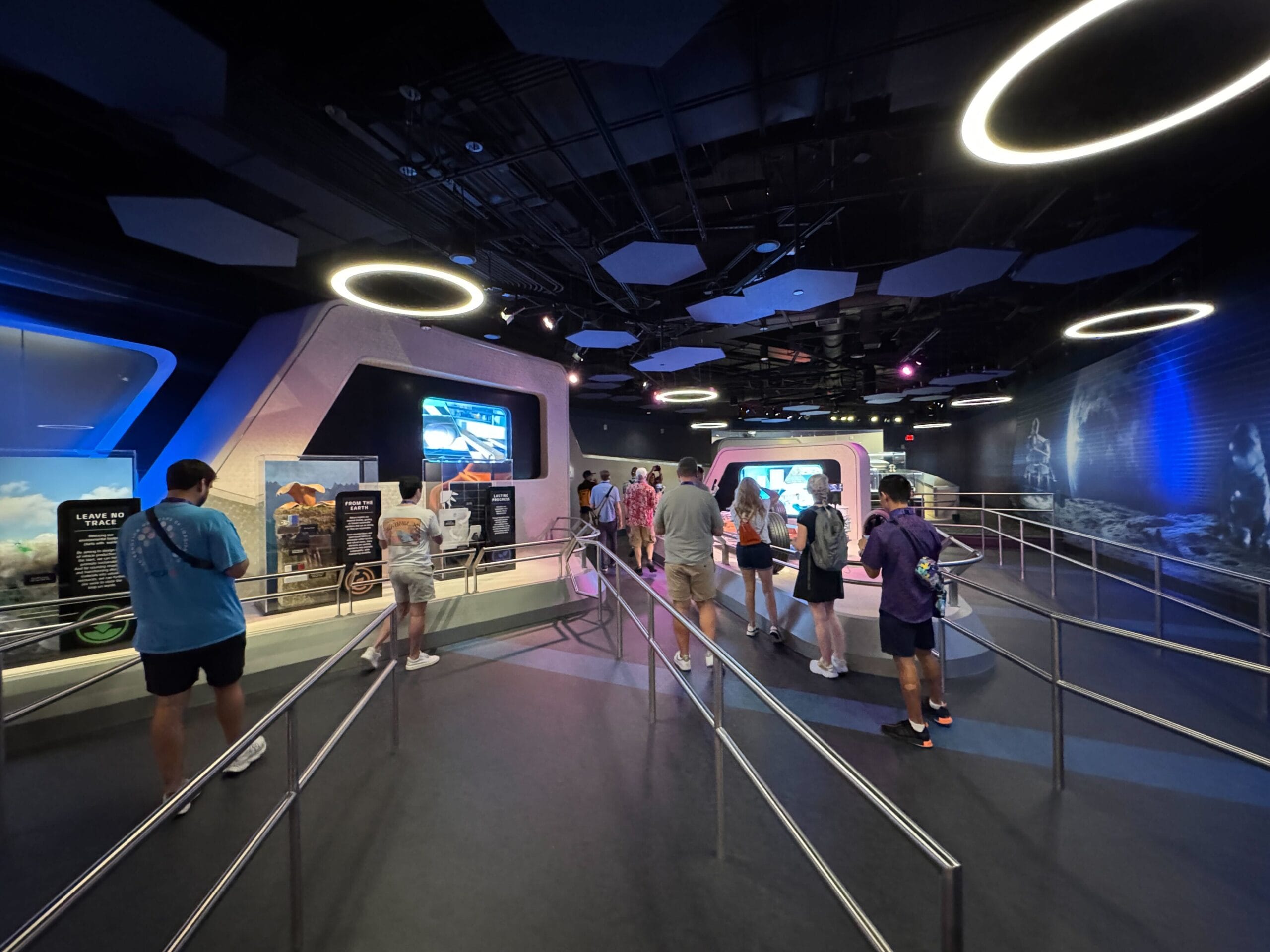 People wait in line inside a futuristic Test Track exhibit with display screens and circular ceiling lights.