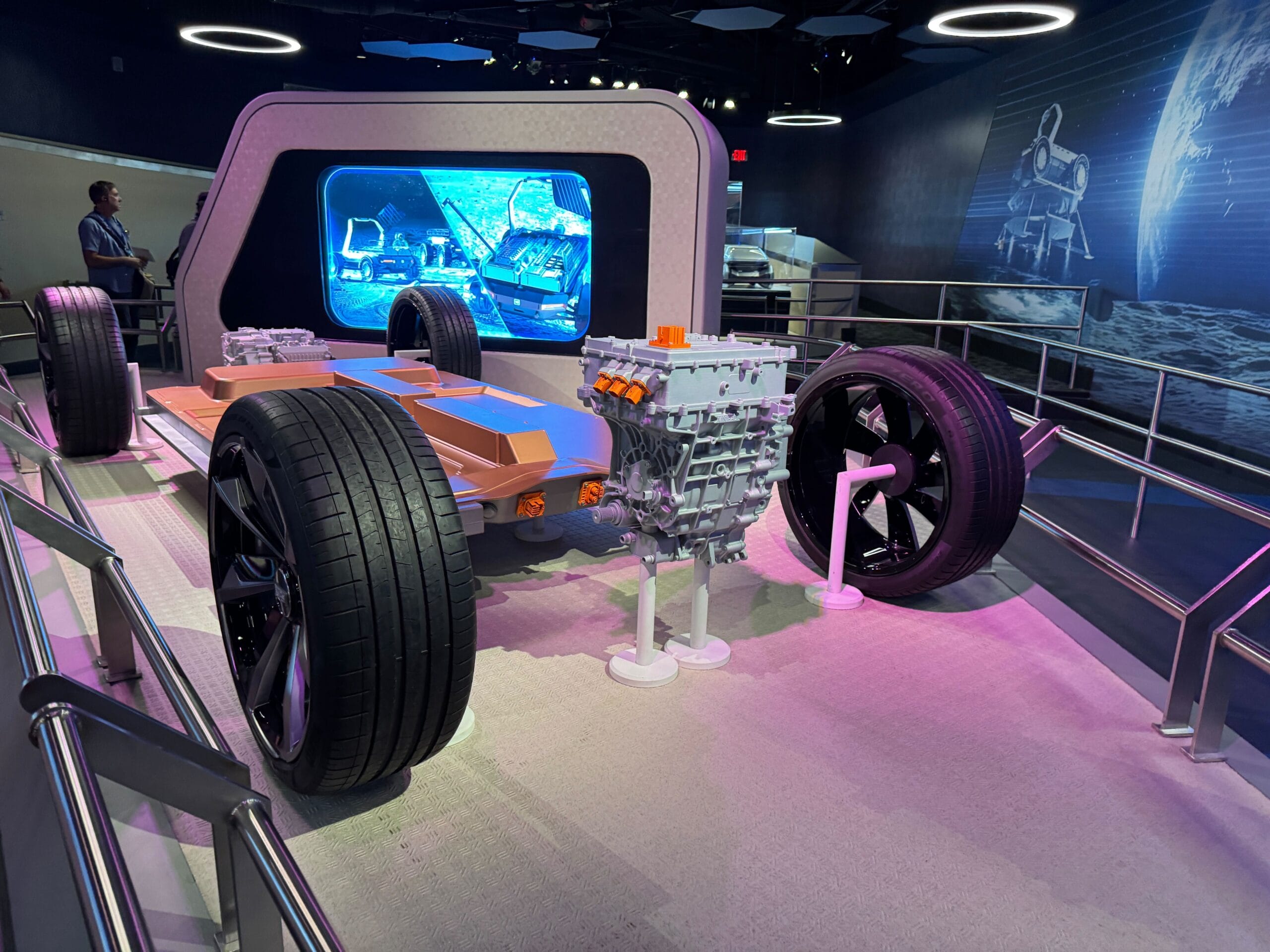 Electric vehicle chassis display with large tires and battery pack, showcased beside a digital Test Track in a modern exhibition space.