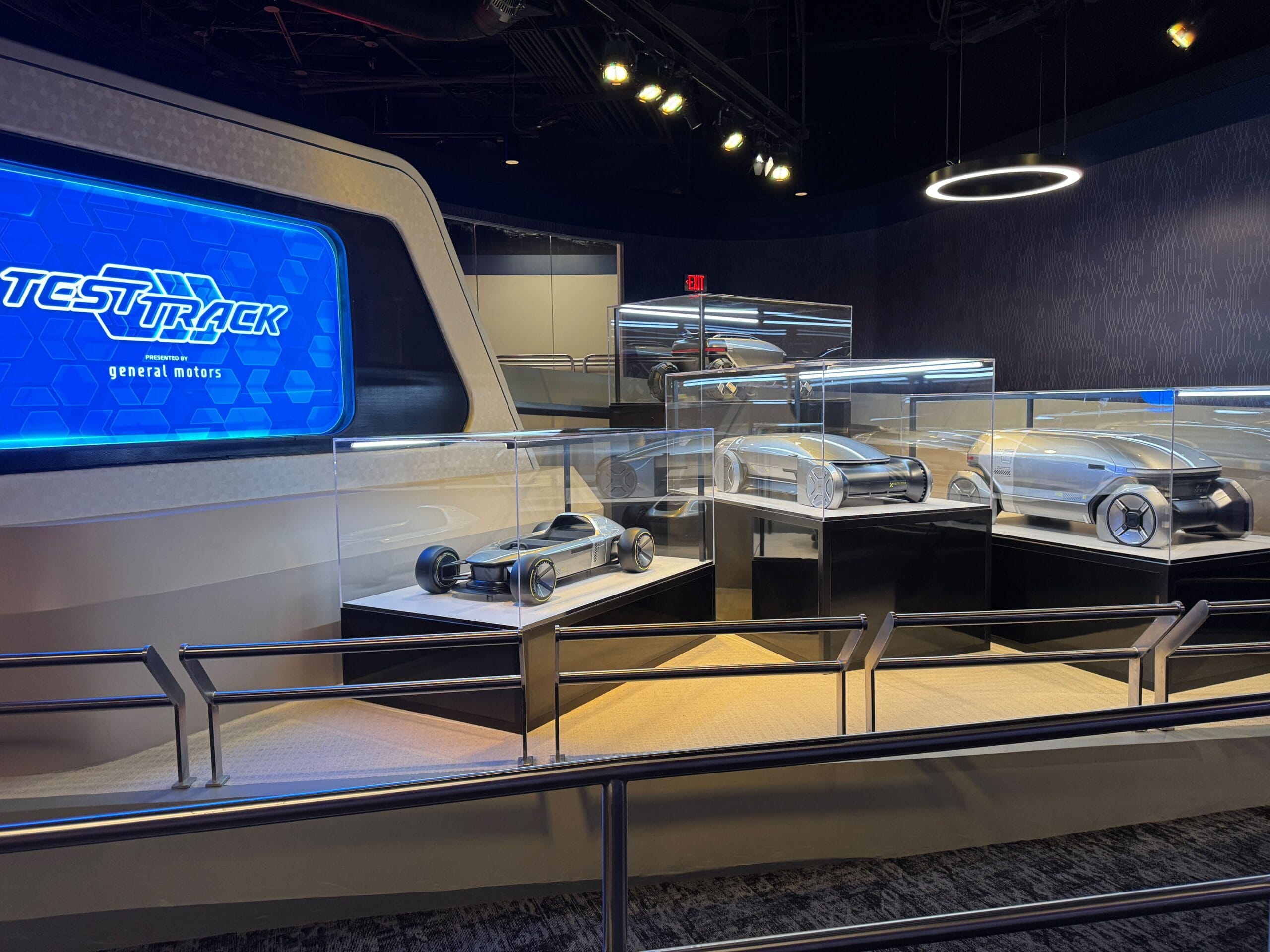Futuristic car models in glass cases at Test Track, with a sign and screens glowing in the background.