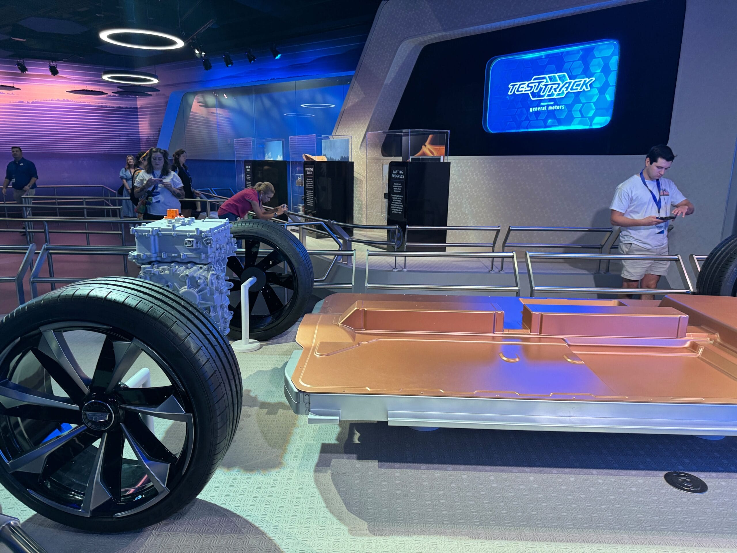 A Test Track car chassis display with large tires and engine, drawing people walking and watching nearby.
