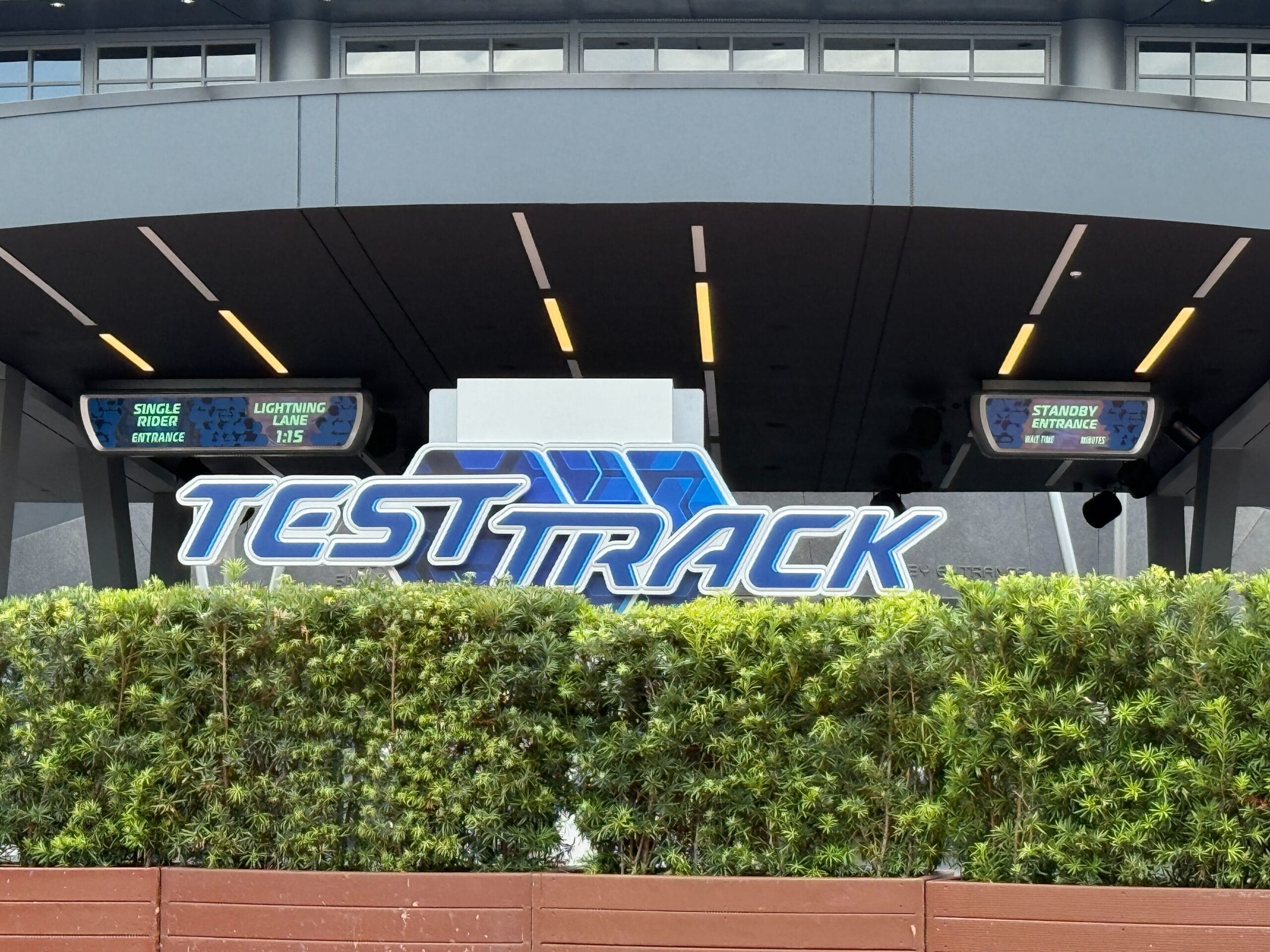 Test Track reopening sign at Epcot with updated wait times above and lush greenery in the foreground. Test Track reopening date sign at Epcot with lightning lane, standby and single rider