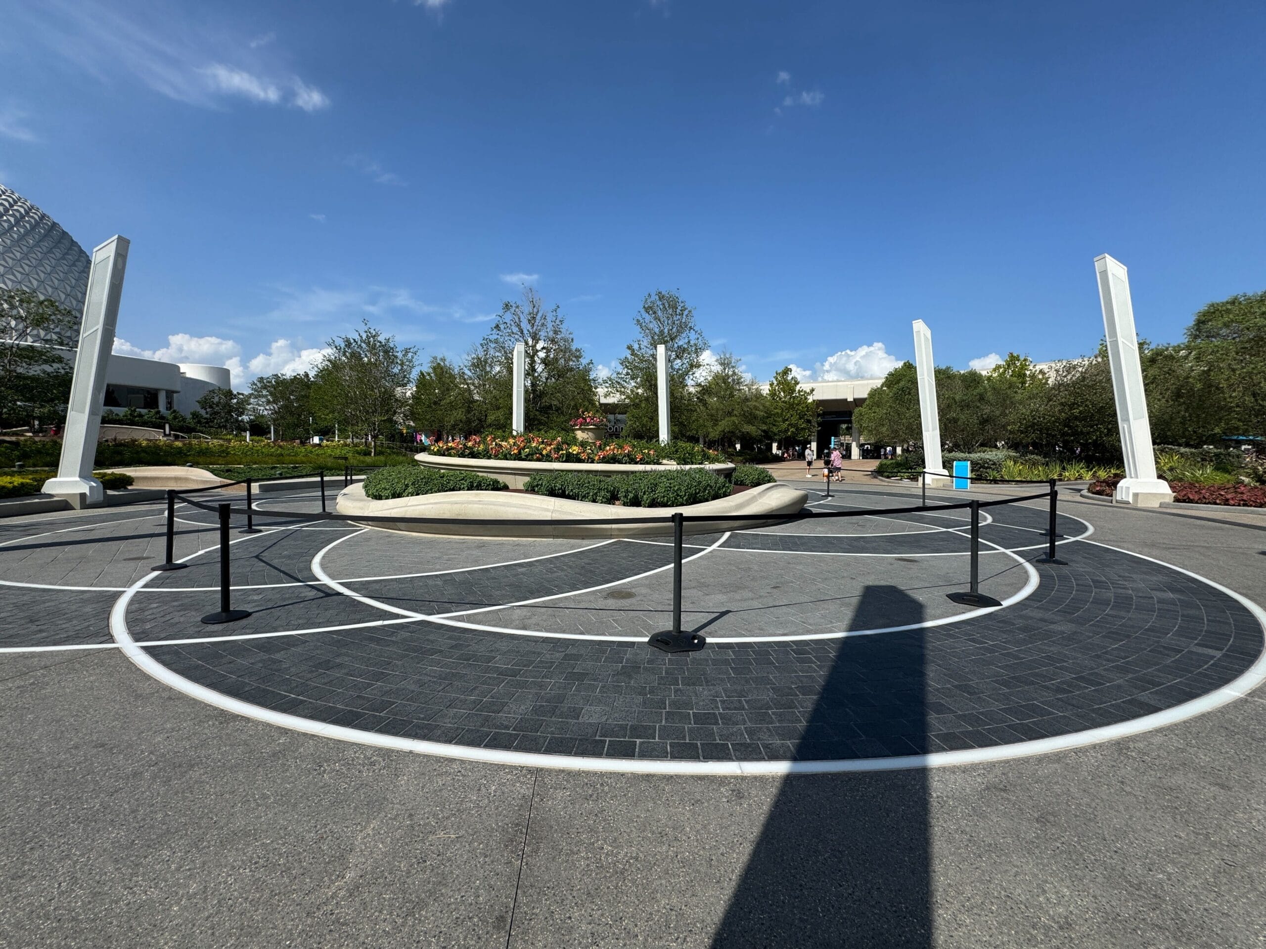 A circular paved area with white lines, pillars, greenery, and a clear blue sky in a public outdoor space.