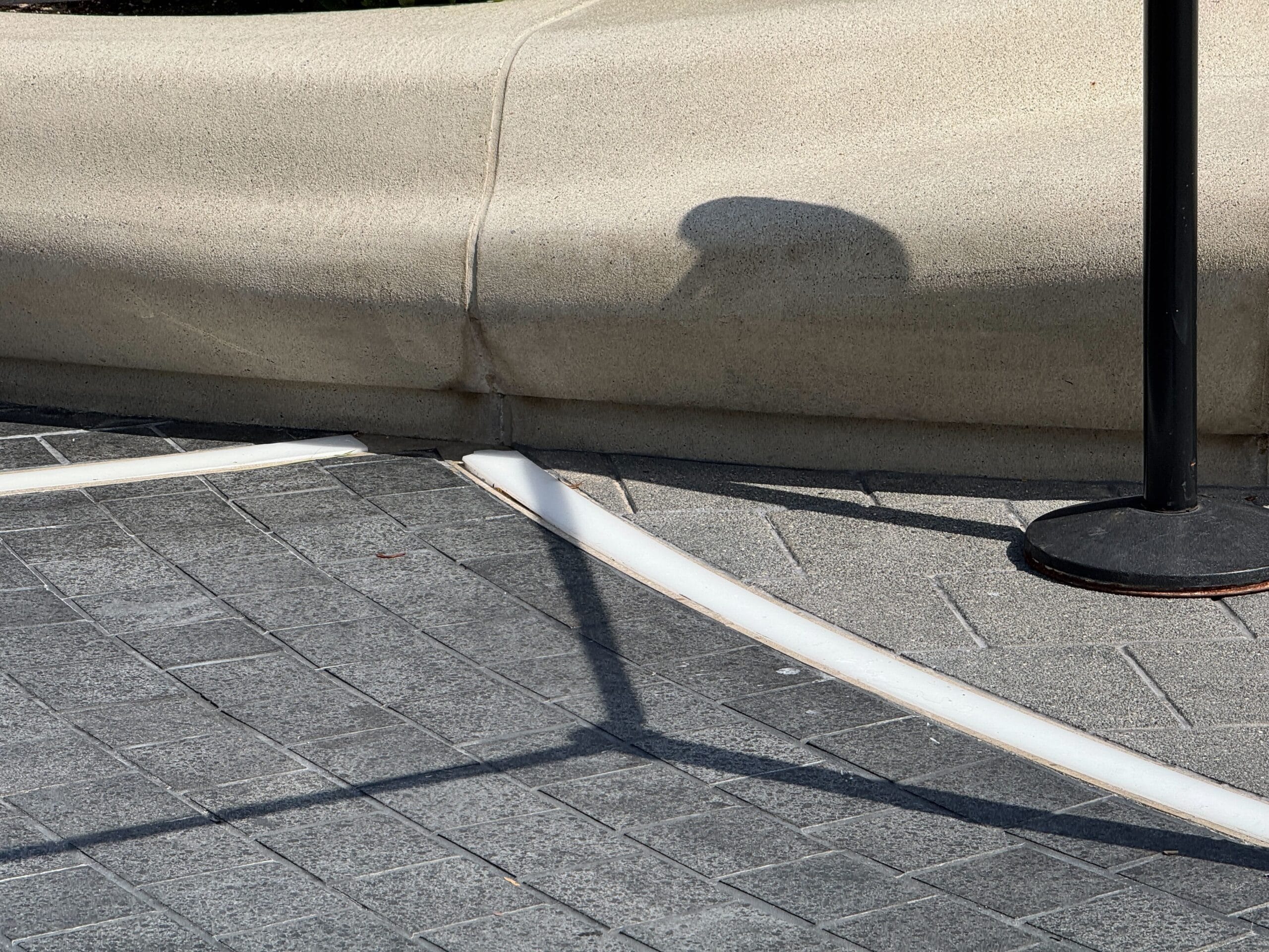 A black pole casts a shadow on World Celebration’s gray Light-Up Pavement near a beige curb with white edging.