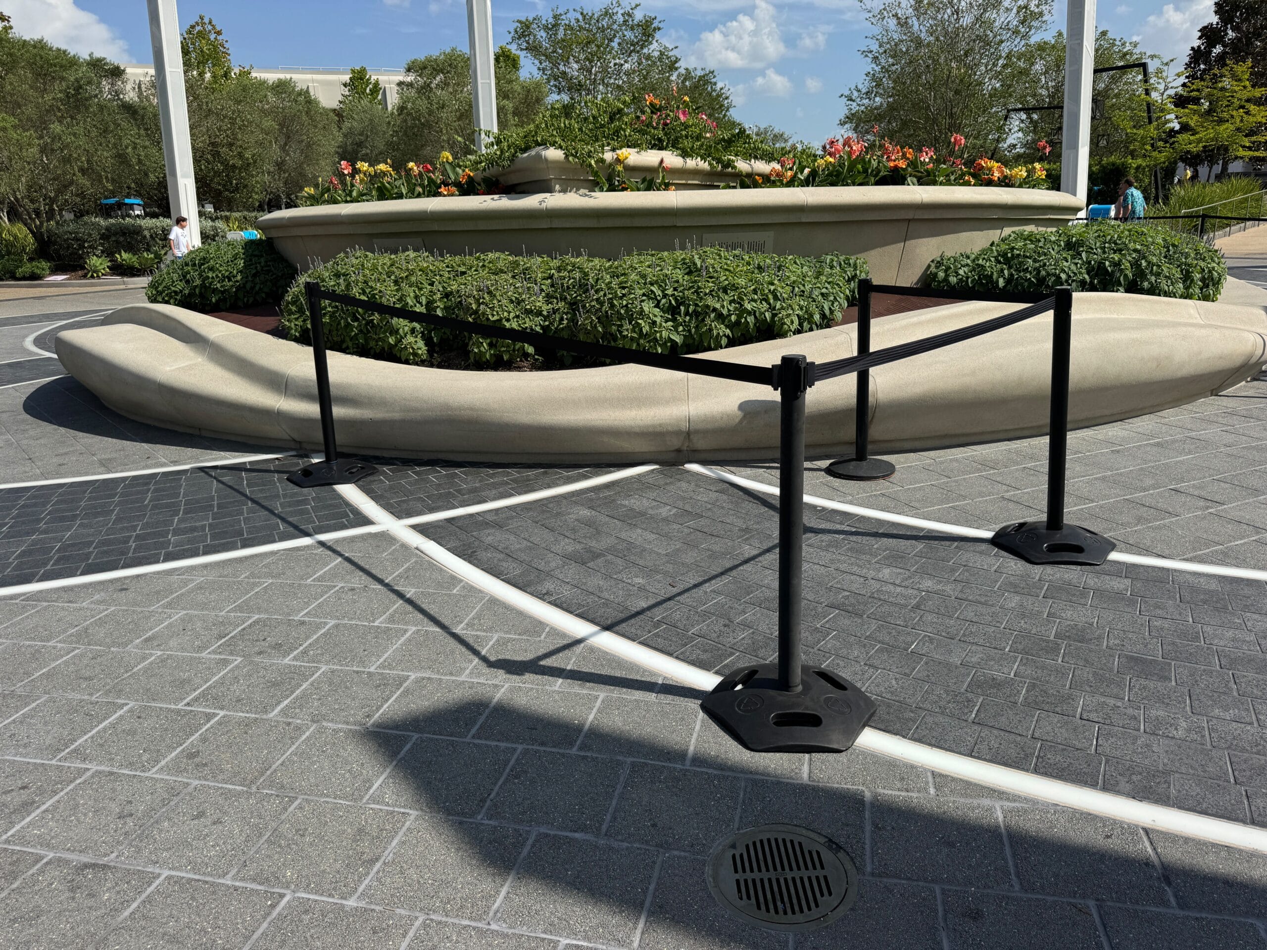 A stone planter with flowers is surrounded by black stanchions and pavement under a sunny sky.
