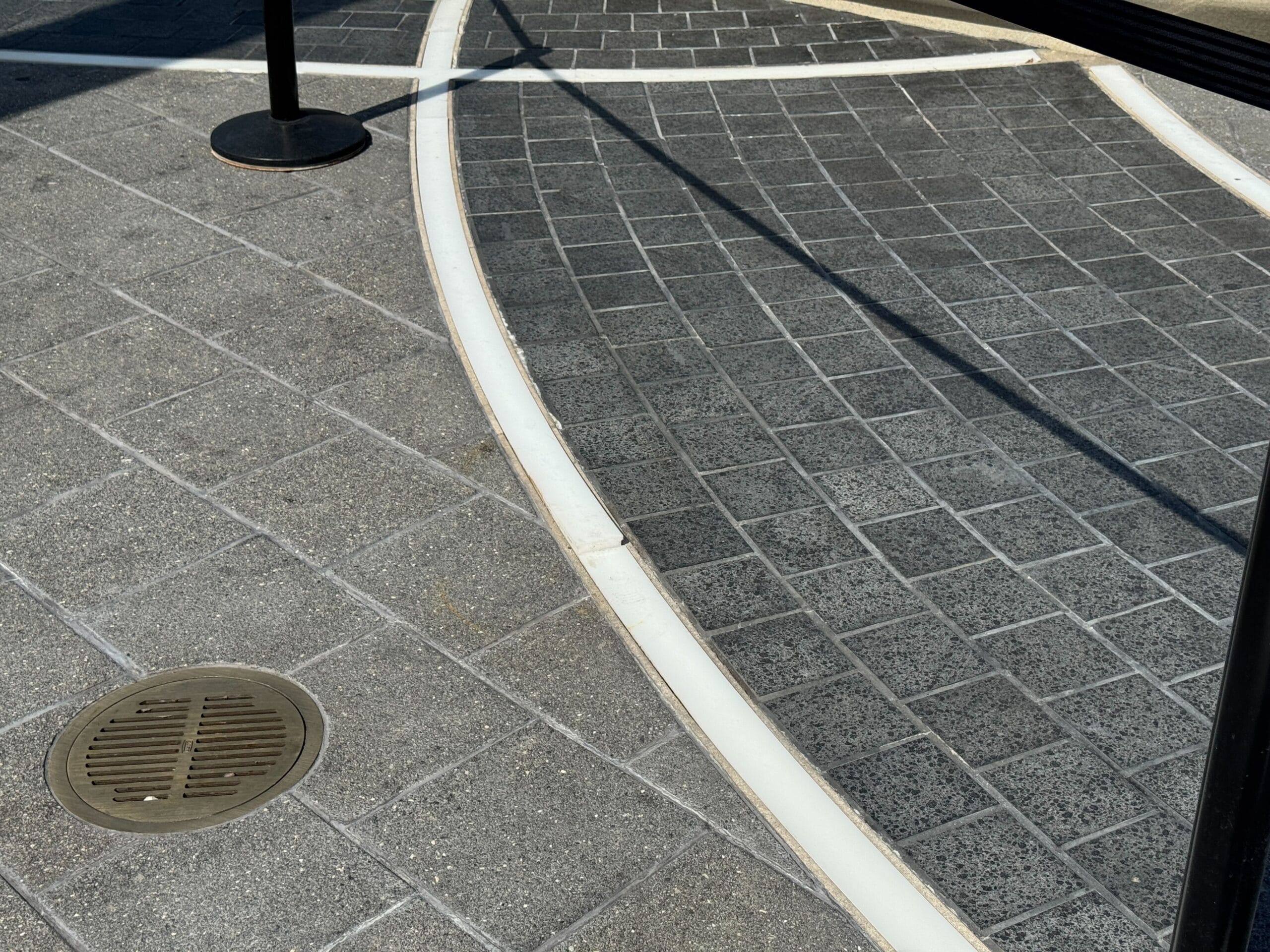 Curved brick and tile Light-Up Pavement with a drain cover and black stanchion casting a shadow, EPCOT World Celebration.